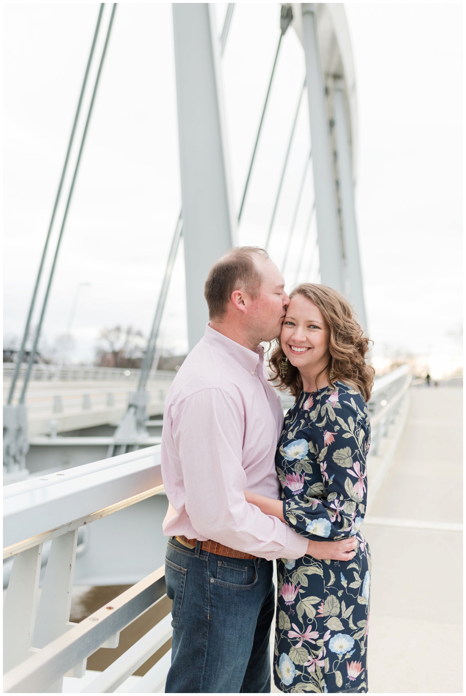 engaged couple stands on bridge at scioto mile. The man is in a pink shirt kissing the girl in a pink and navy floral dress. She is smiling at the camera 