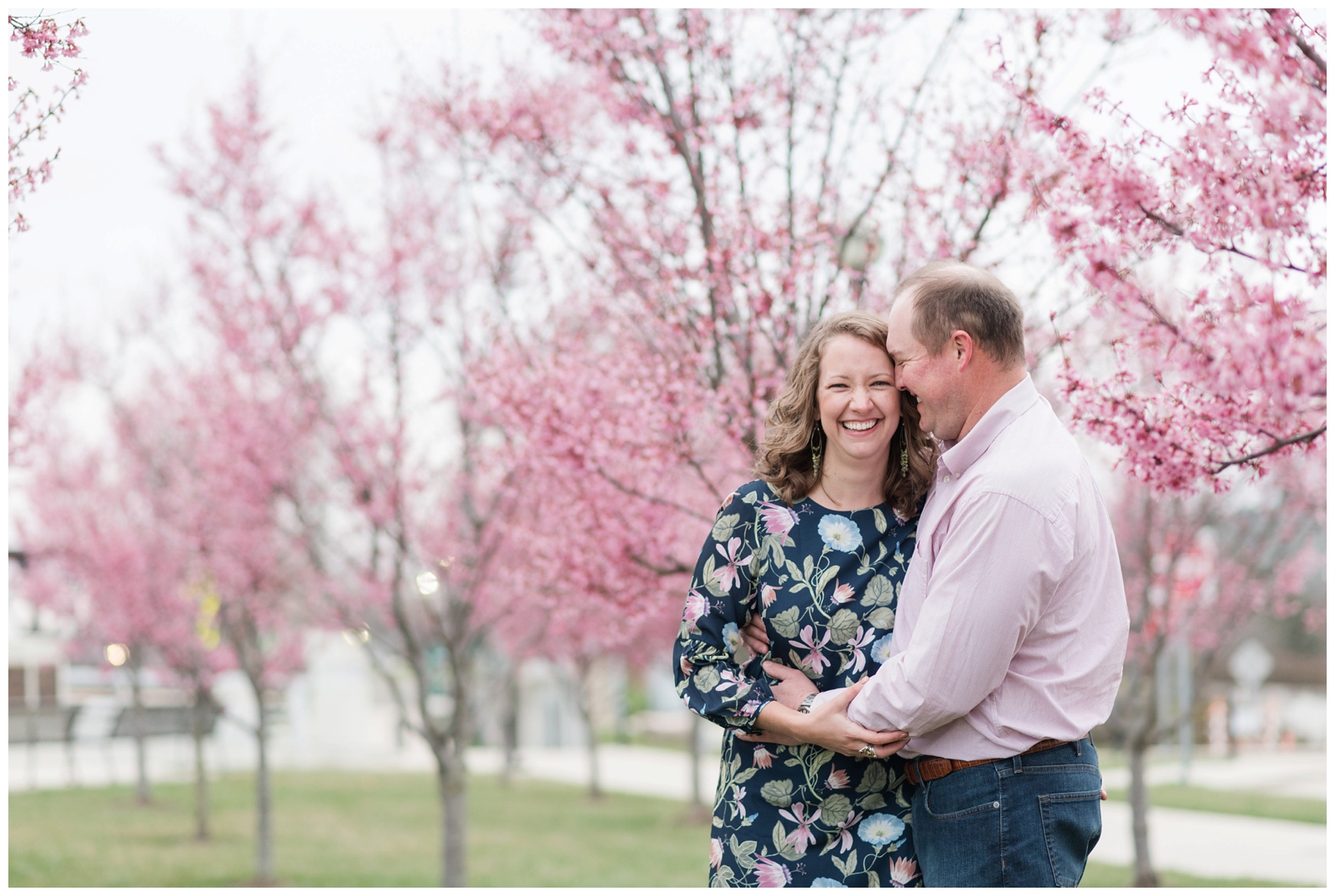 Cheerful couple laughing at each other with red bud spring blossoms all around them and the columbus ohio city skyline behind them at scioto mile in downtown columbus by Pipers Photography 