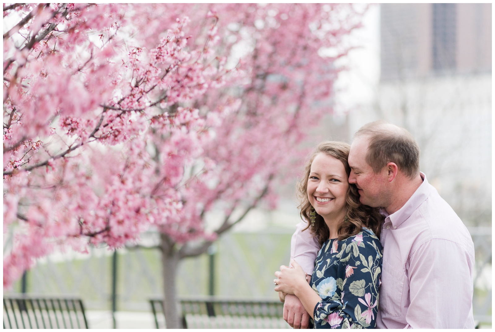Cheerful couple laughing at each other with red bud spring blossoms all around them and the columbus ohio city skyline behind them at scioto mile in downtown columbus surrounded by red bud trees Scioto Mile Engagement Session Columbus, Ohio