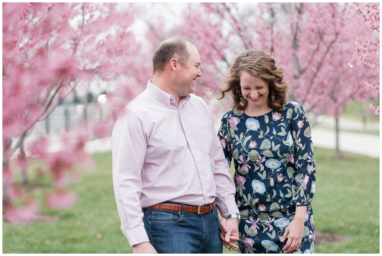 Cheerful couple laughing at each other and walking towards the camera with red bud spring blossoms all around them and the columbus ohio city skyline behind them at scioto mile in downtown Columbus Scioto Mile Engagement Session Columbus, Ohio