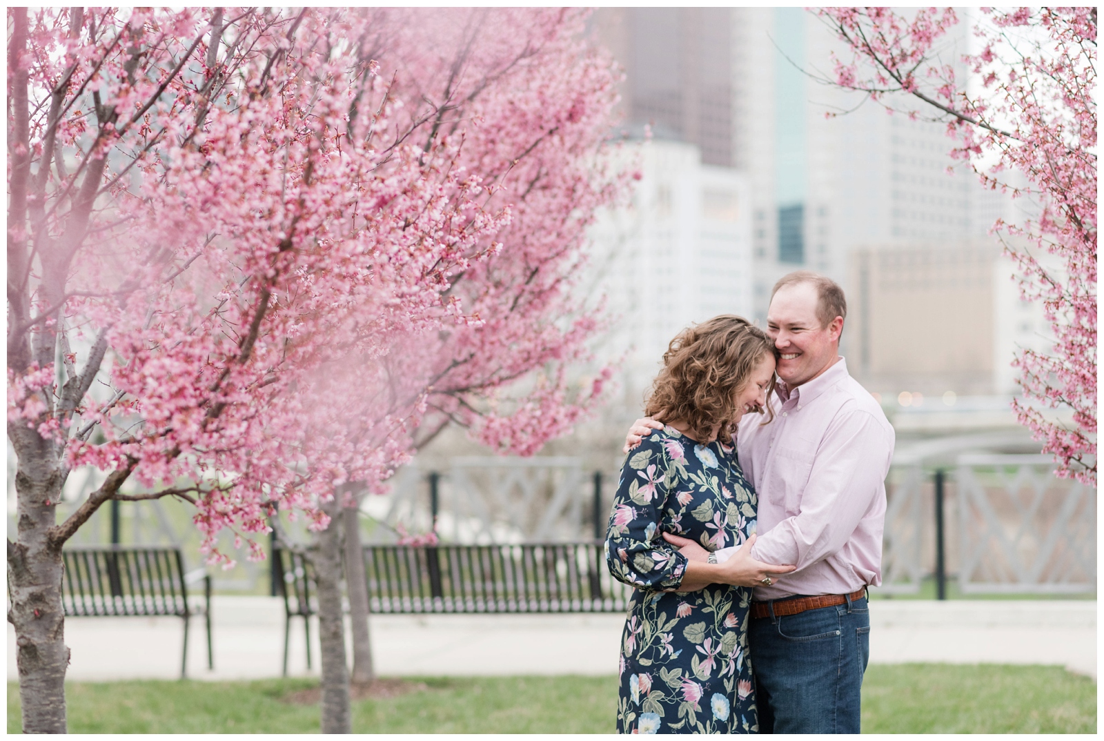 Cheerful couple laughing at each other with red bud spring blossoms all around them and the columbus ohio city skyline behind them at scioto mile in downtown columbus 