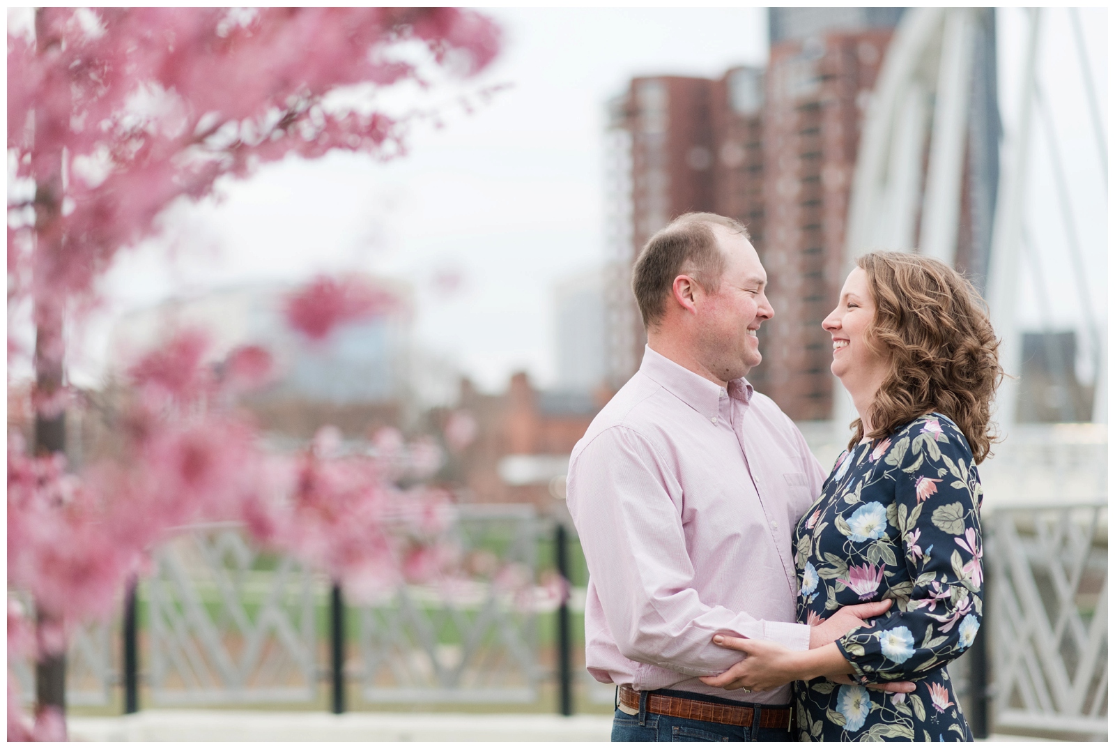 an engaged couple looks at each other with a columbus ohio bridge and skyline behind them and spring red bud trees surrounding them . Scioto Mile Engagement Session Columbus, Ohio