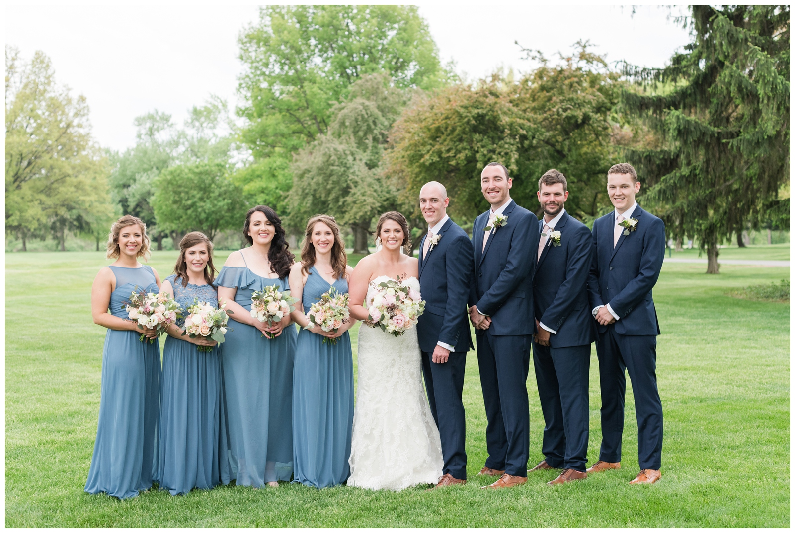 bridesmaids in blue gowns stand next to bride while groomsmen in navy suits stand by groom during wedding portraits in Ohio