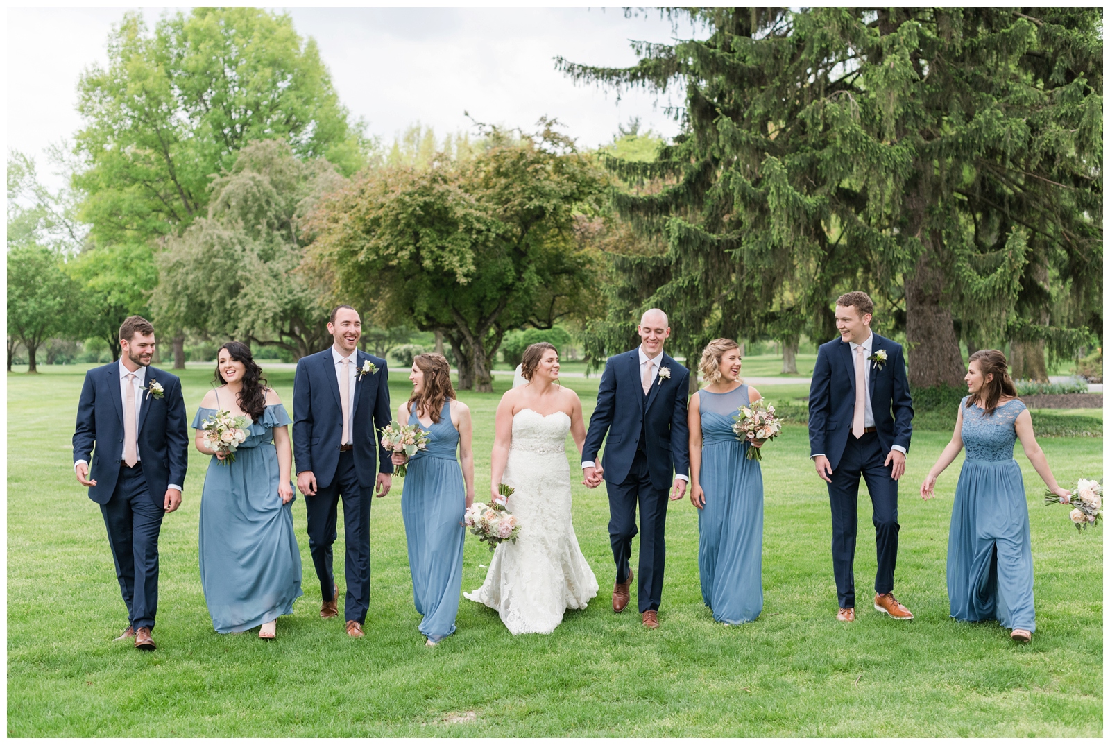 bride and groom walk beside bridesmaids in blue gowns and groomsmen in navy suits