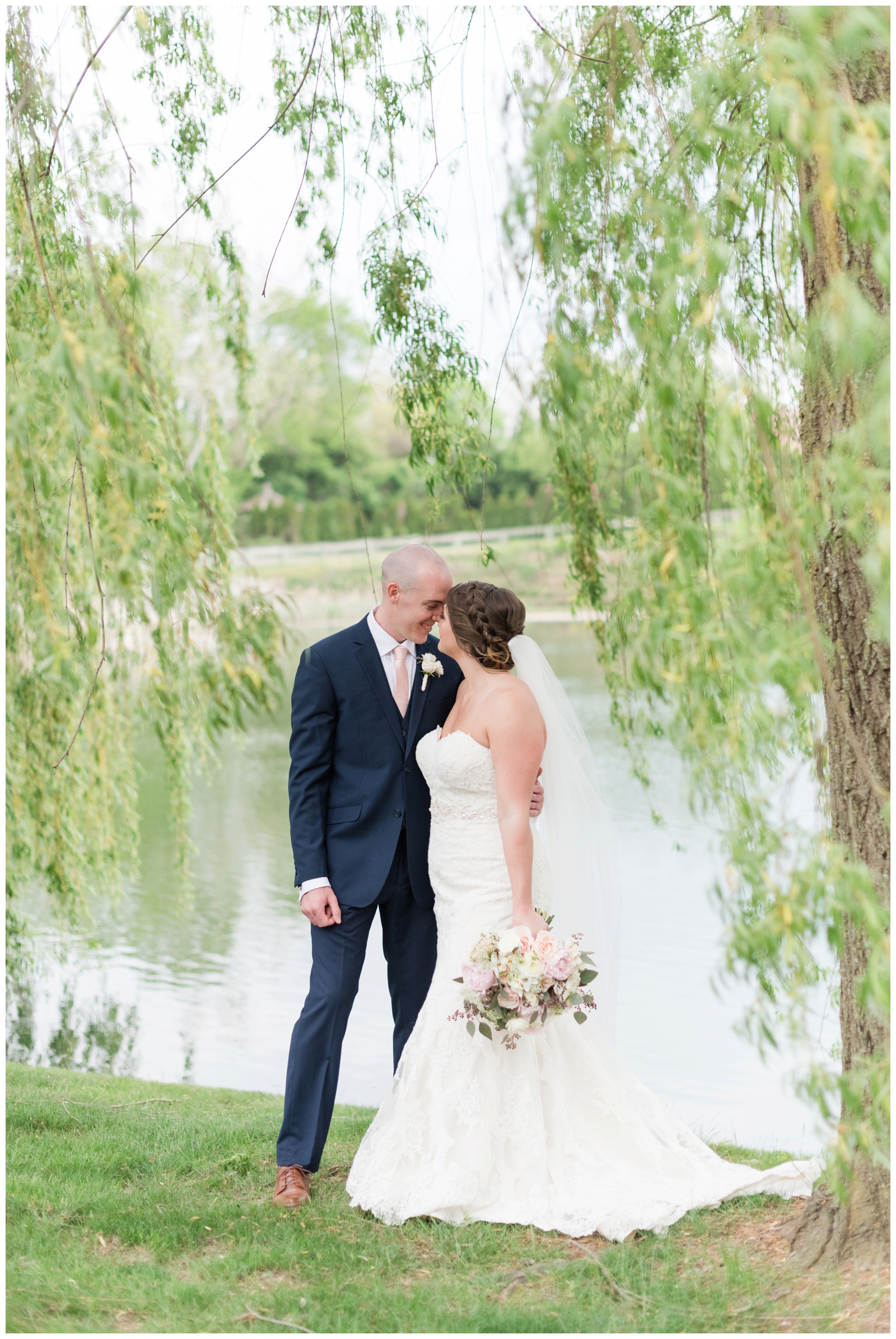 bride and groom nuzzle noses while bride holds wedding bouquet of pink and white flowers under willow tree by lake