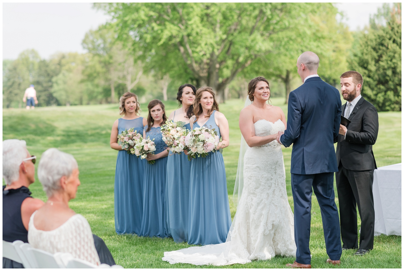  bride and groom exchange vows while family watches with bridesmaids in blue gowns nearby at Brookside Golf and Country Club