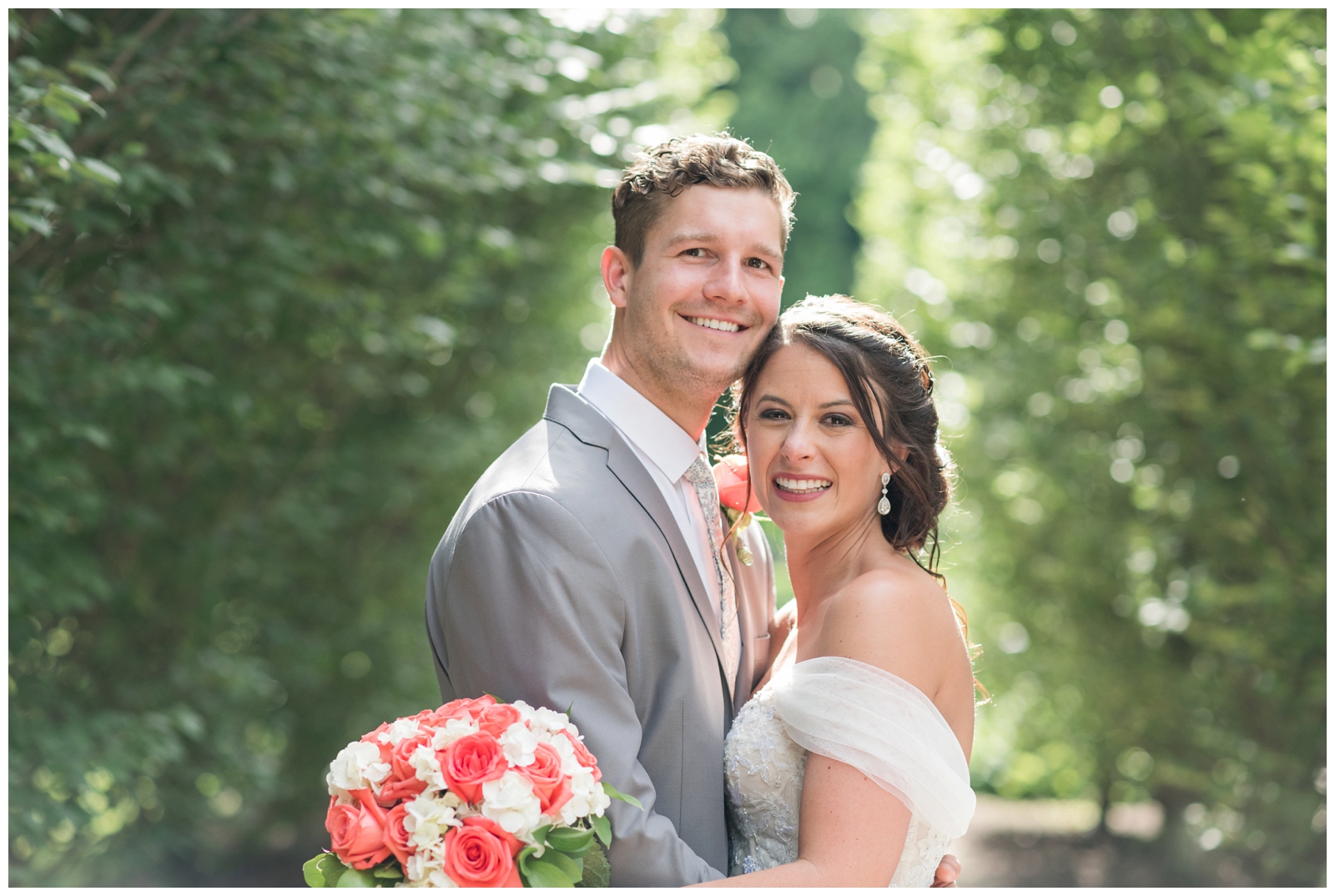 bride hugs groom in grey suit while holding bouquet with white flowers and coral roses