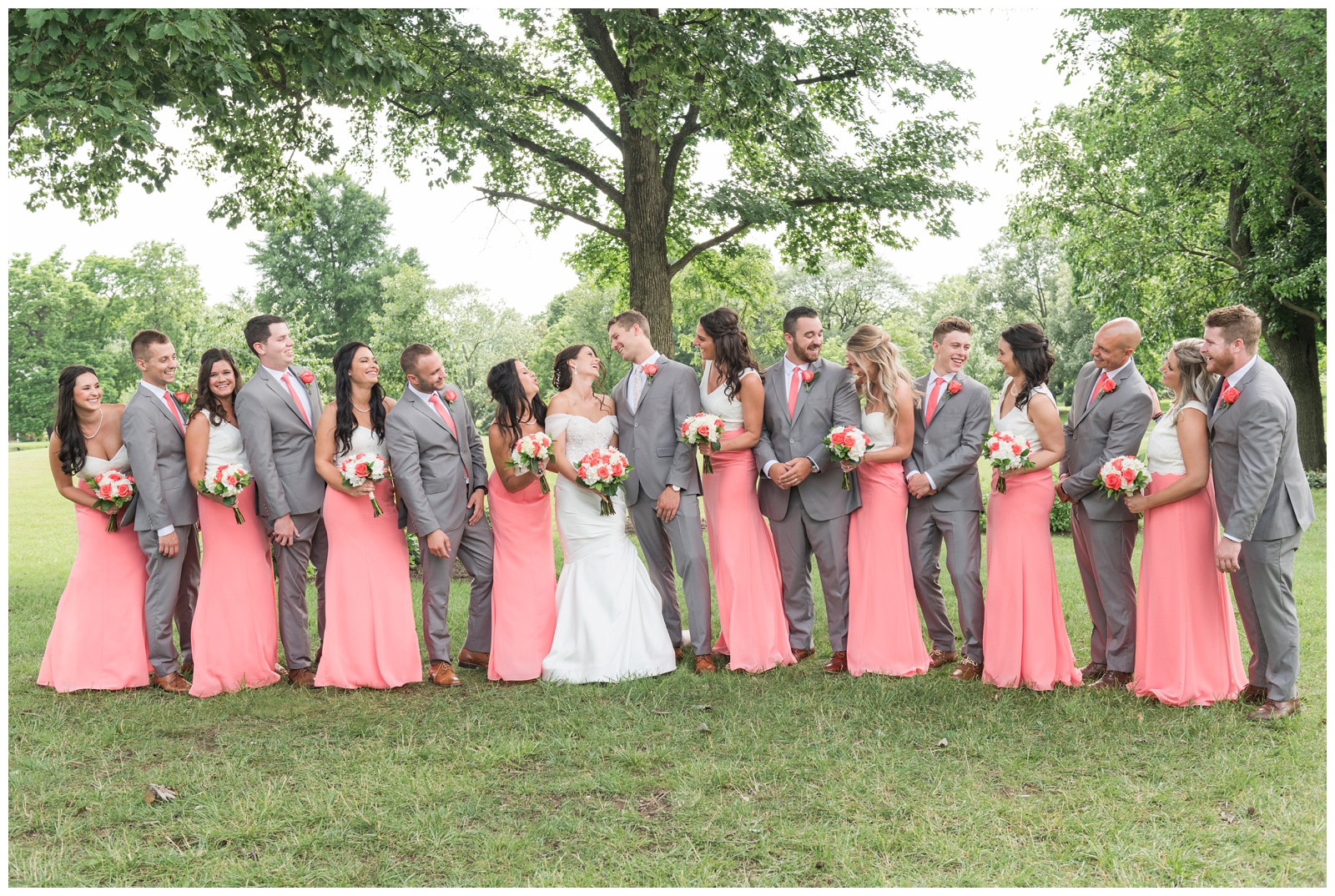 large bridal party in coral and white dresses and grey suits look at bride and groom