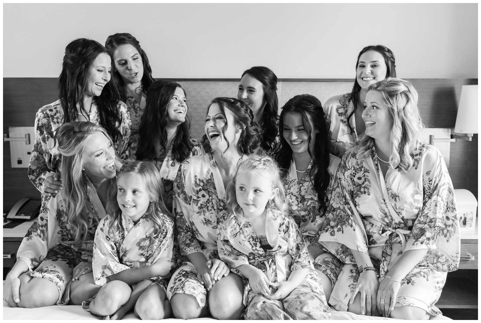 bride laughs with bridesmaids and two flower girls in floral robes on bed before Ohio wedding day