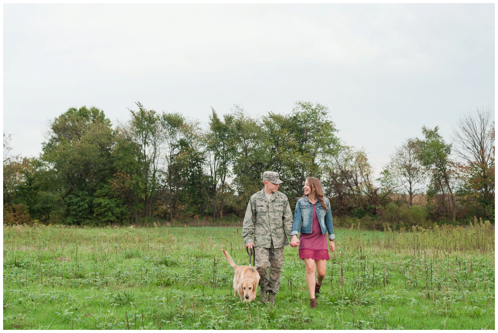 girl and boy walking with their dog through a grass field at Engaged couple looking at camera in wheat field with burgundy dress and military bdus on in Country Engagement Session Sunbury Ohio