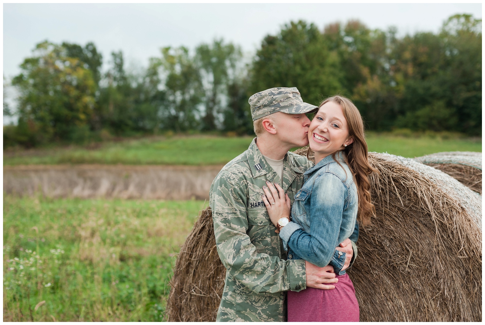 engaged man kissing girl looking at the camera in a grass field with military uniform on and burgundy dress in a country engagement session in sunbury ohio