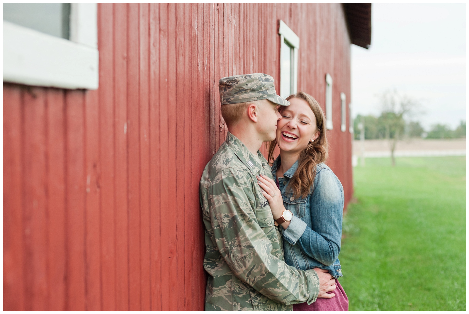 engaged girl laughing at her fiancé during their country engagement session leaning against a red barn with military uniform on and burgundy lace dress 