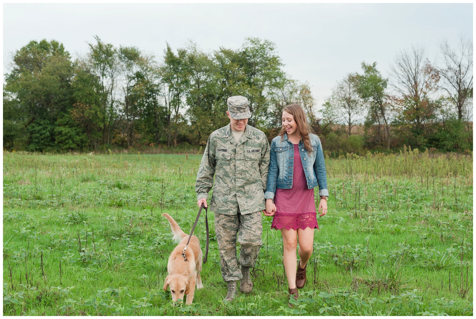 Engaged couple walking through field with burgundy dress and military outfit with their golden lab dog on in Country Engagement Session Sunbury Ohio