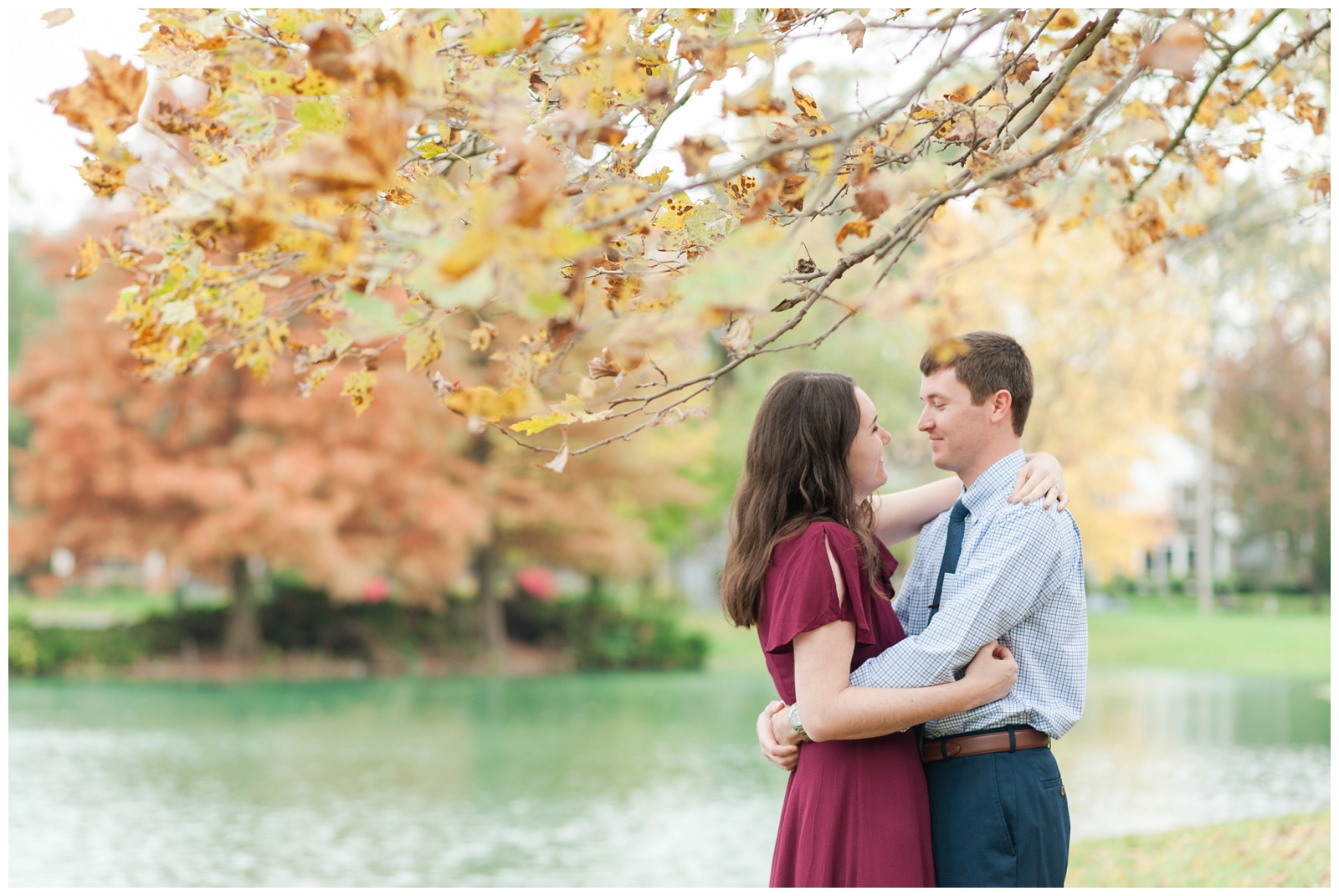 bride and groom looking at each other for their engagement photos under a fall colored tree with a pond behind them 
