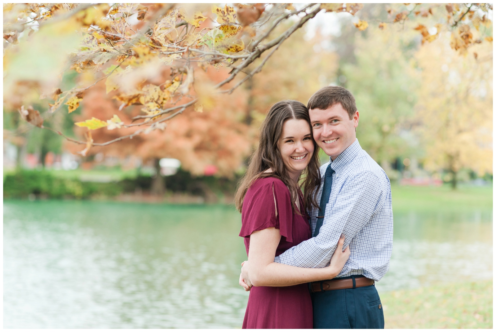 engaged couple looking at the camera with a pond and beautiful fall leaves behind them during their engagement session in a warm embrace 