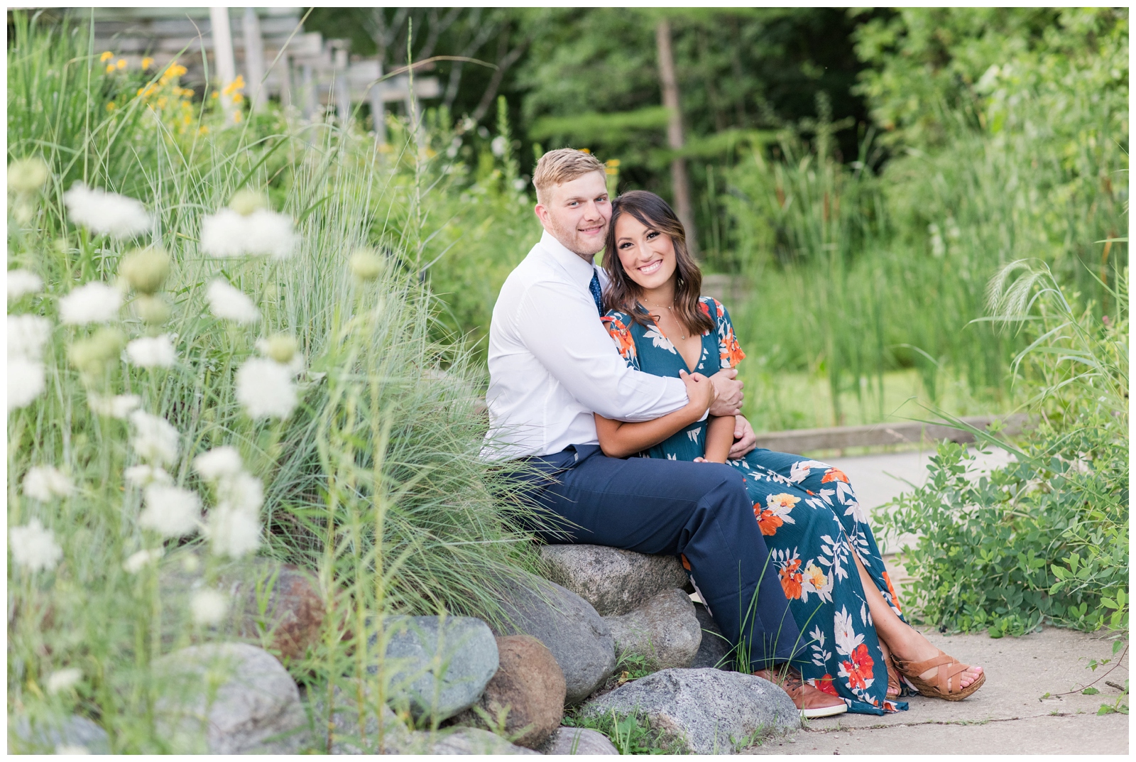engaged couple happily smiling at the camera in their formal wear while sitting on a rock surrounding by green and white greenery and flowers in a garden during a summer evening 