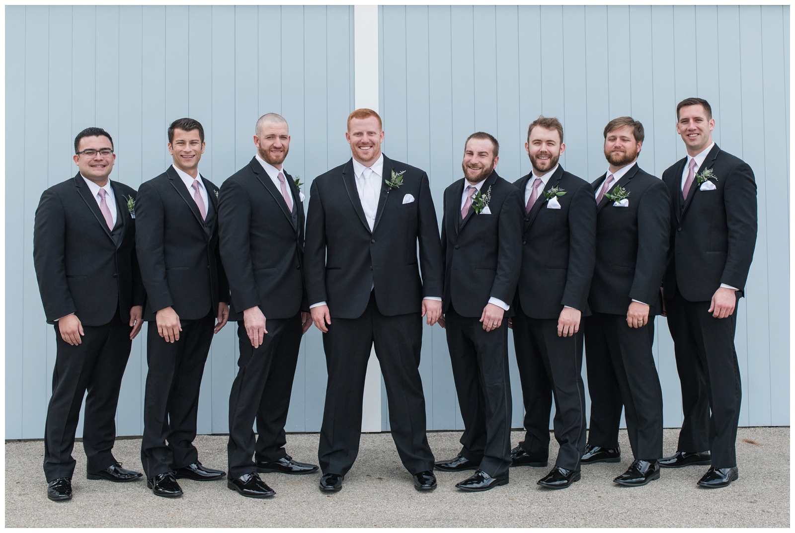 groomsmen in black suits pose with groom by blue wall at Pretty Prairie Farms