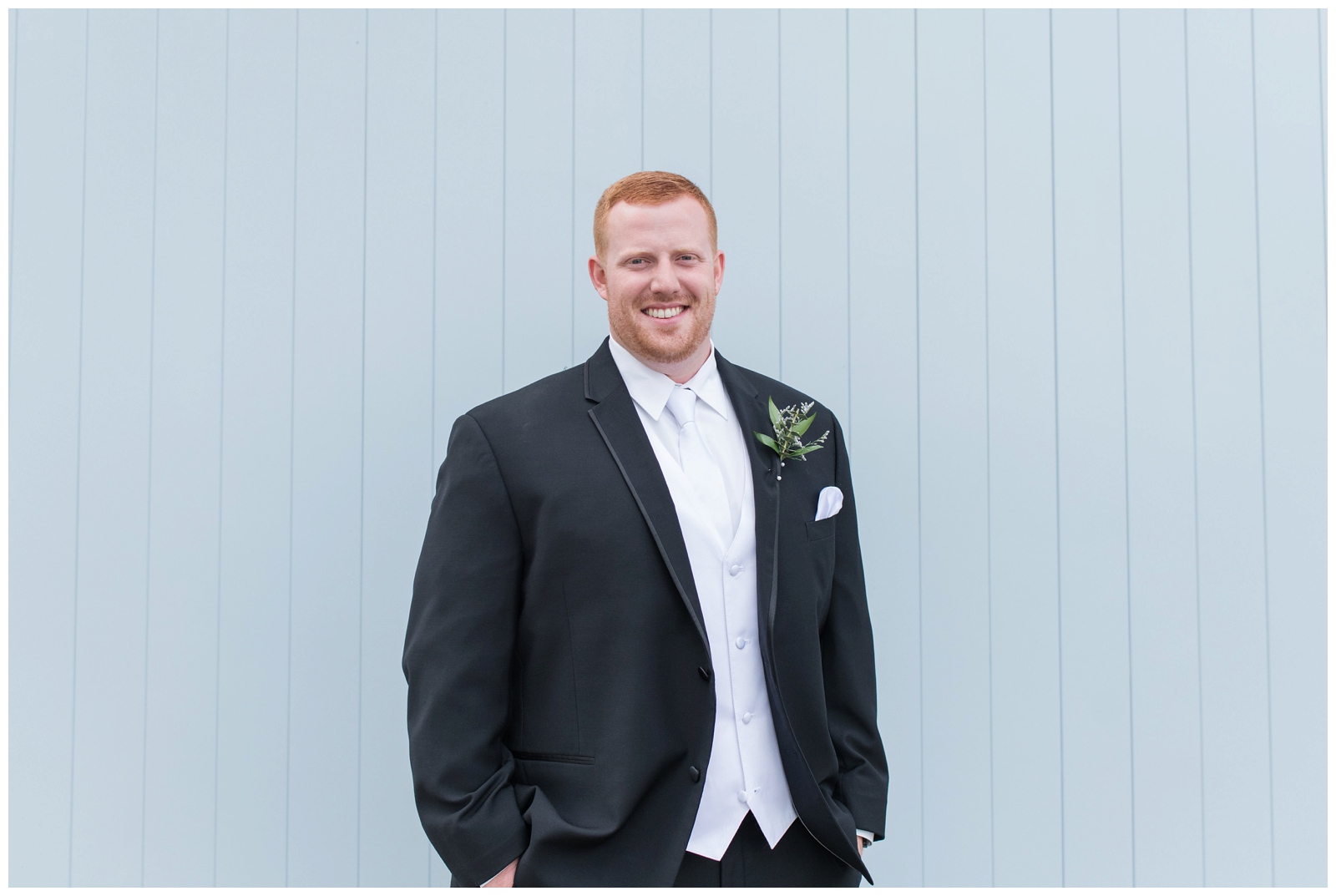 groom smiles at camera and poses by blue wall at Pretty Prairie Farms during Ohio wedding day