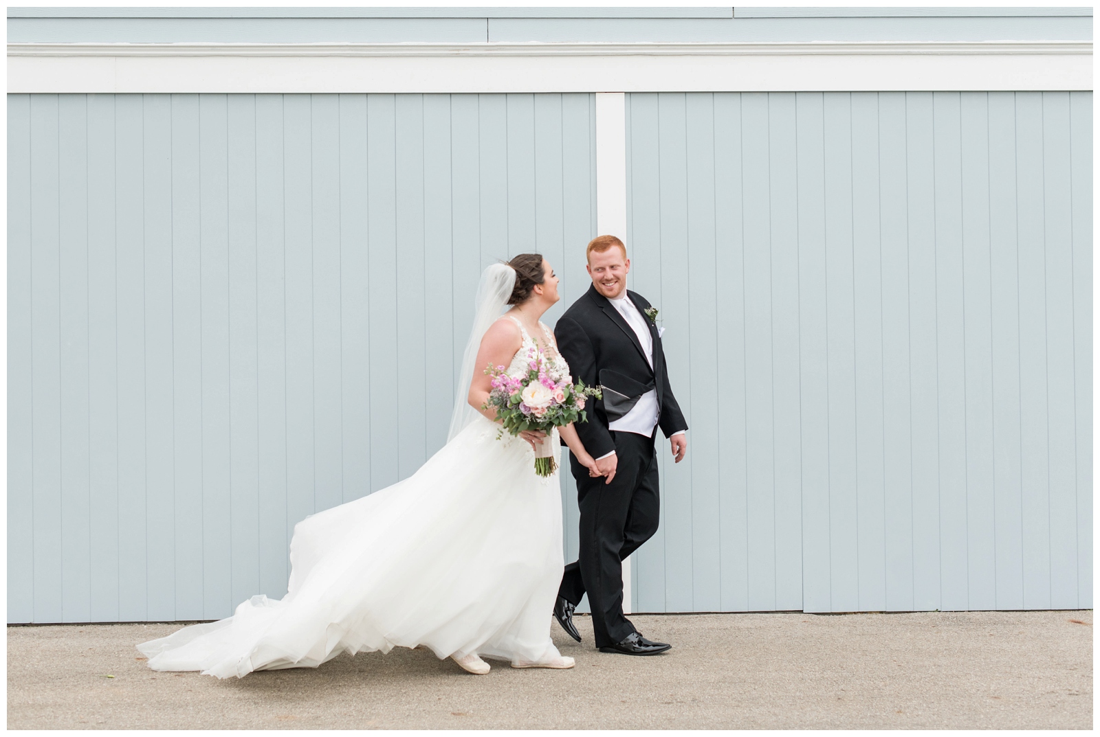 Bride and Groom are walking across a pale blue barn wall during their wedding portraits at Pretty Prairie Farm Weddings in Urbana, Ohio.