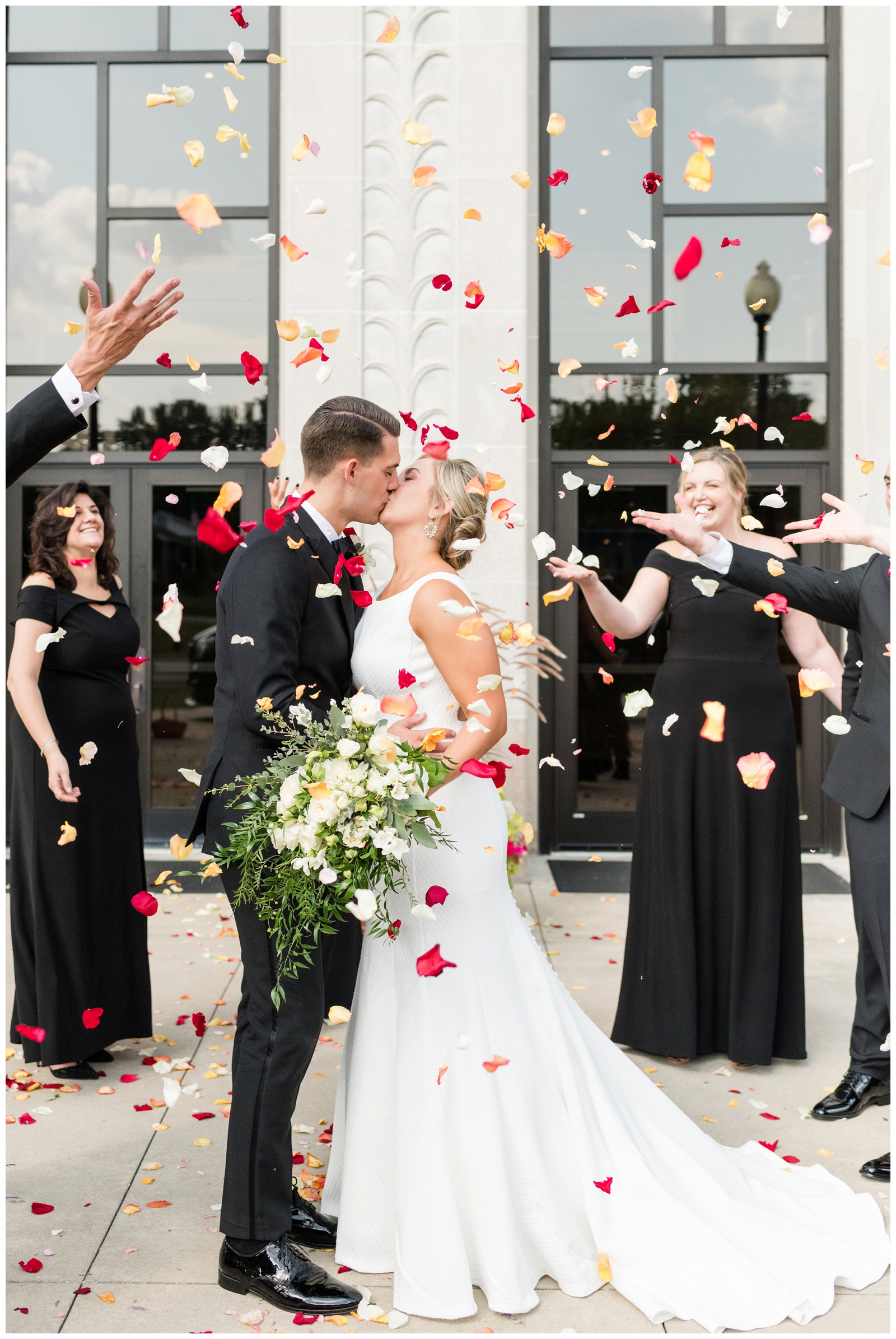bride and groom kiss while red and orange flower petals are thrown around them by bridal party