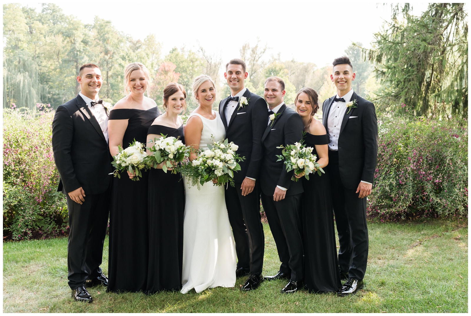 bridesmaids in black gowns and groomsmen in tuxes surround bride and groom at Gervasi Vineyard