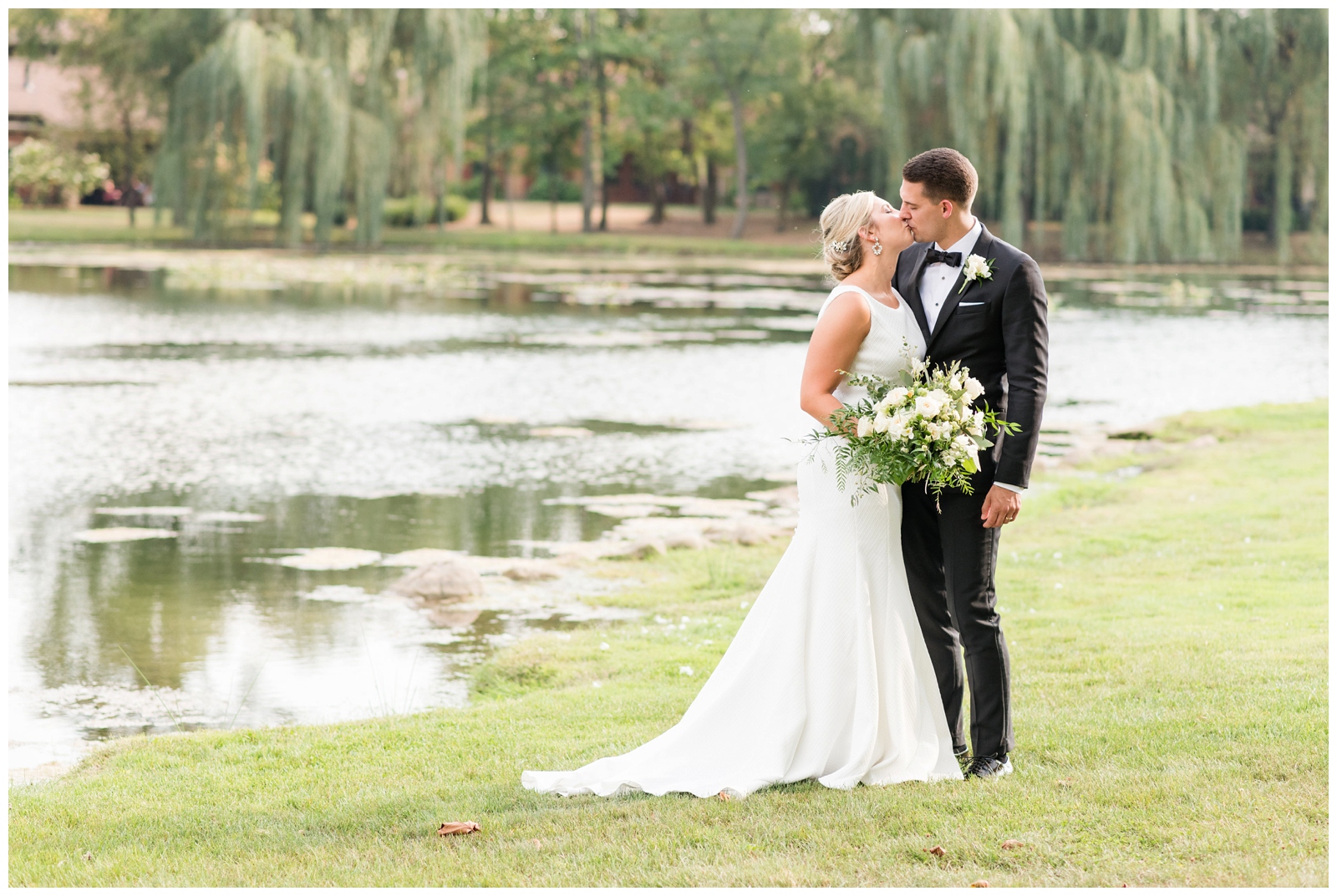 bride and groom kiss by water at Gervasi Vineyard while bride holds all-white wedding bouquet