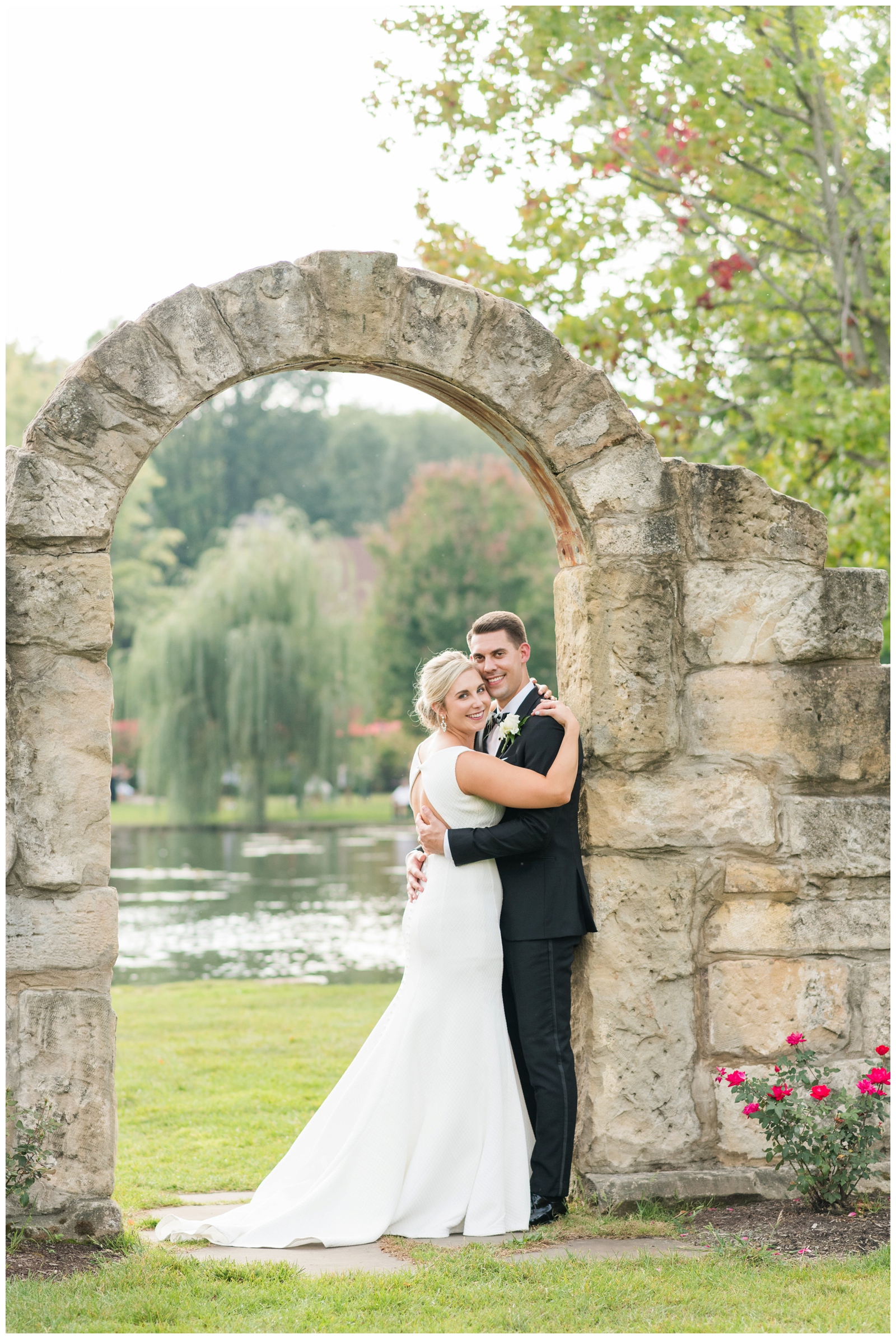 bride and groom hug under stone archway in front of pond at Gervasi Vineyard in Canton OH