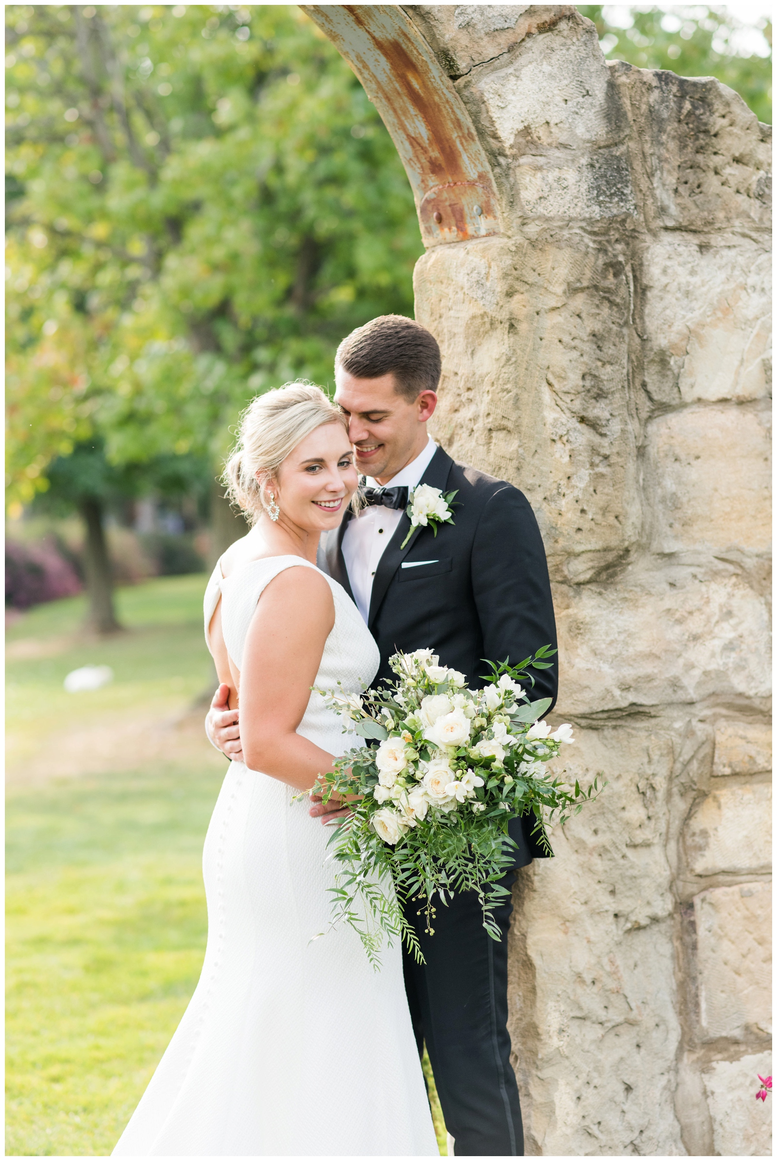 groom looks at bride while she holds all-white bouquet under stone archway at Gervasi Vineyard