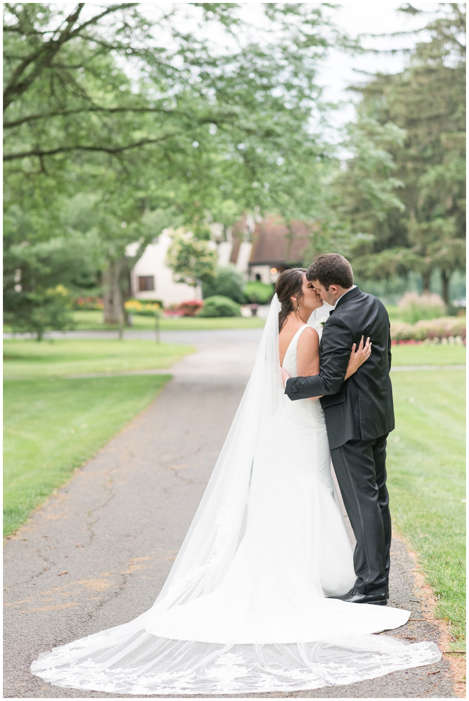 newlyweds kiss along driveway at Brookside Golf and Country Club while bride's lace veil is laid out behind them