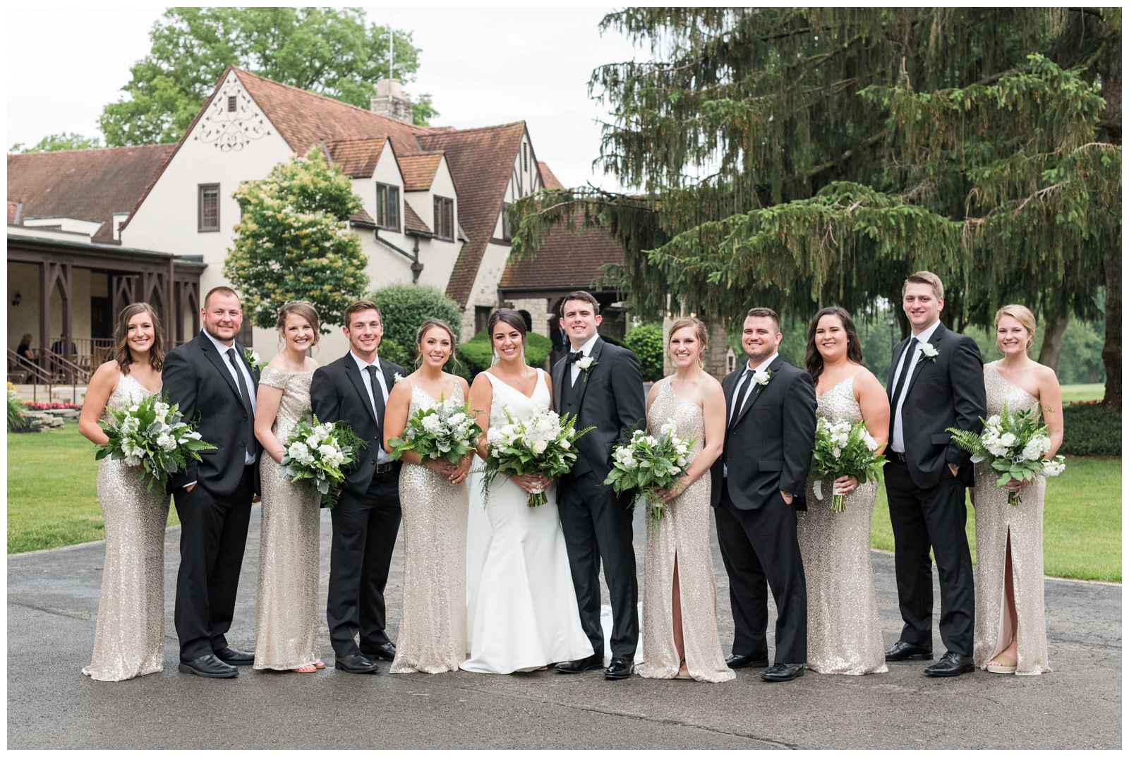 bridal party in gold gowns and black suits pose with bride and groom in front of Brookside Golf and Country Club 