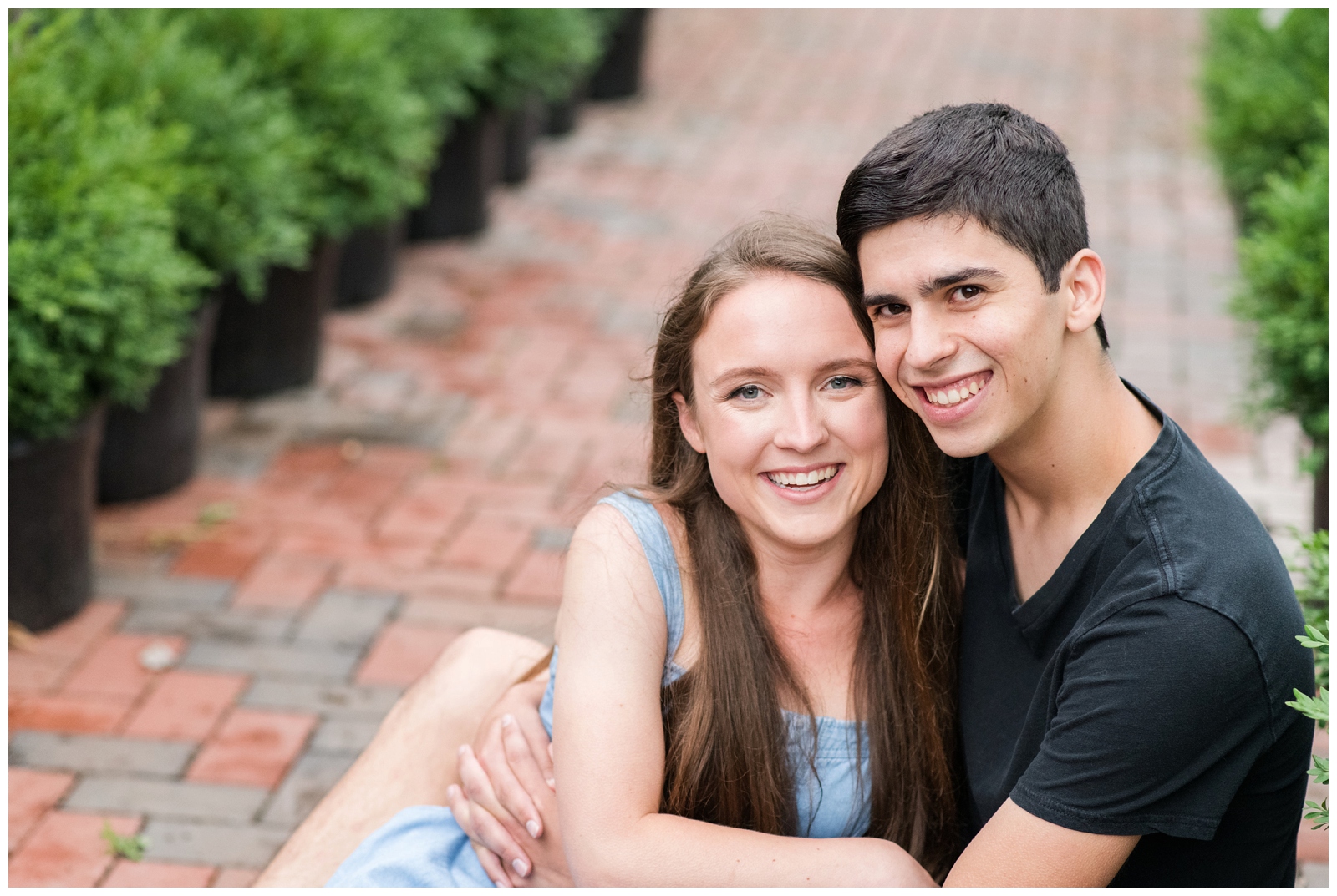 engaged couple sits on brick walkway with green plants around them during greenhouse inspired engagement session