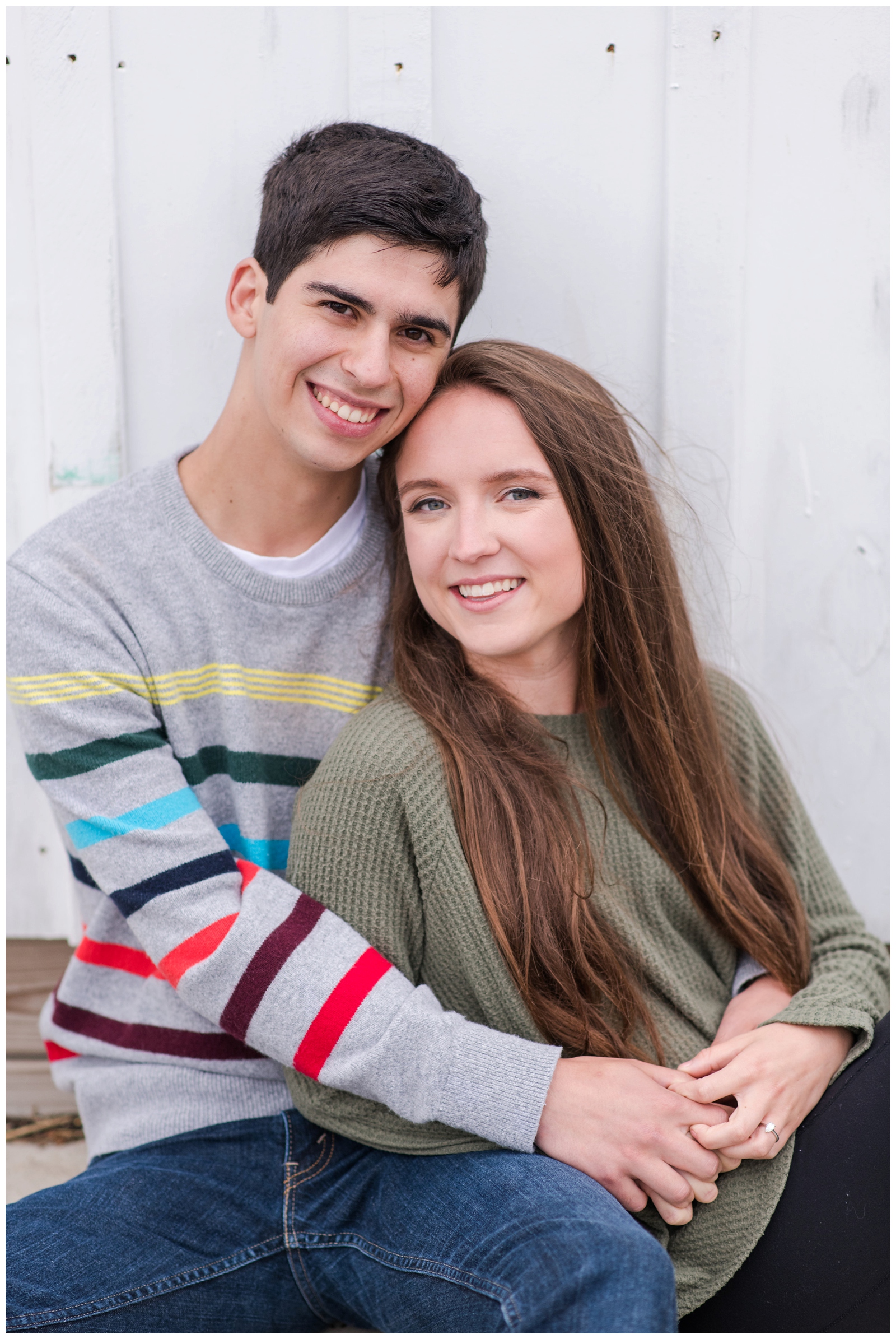 young couple snuggles in cozy sweaters along white wall at Oakland Nursery for engagement portraits