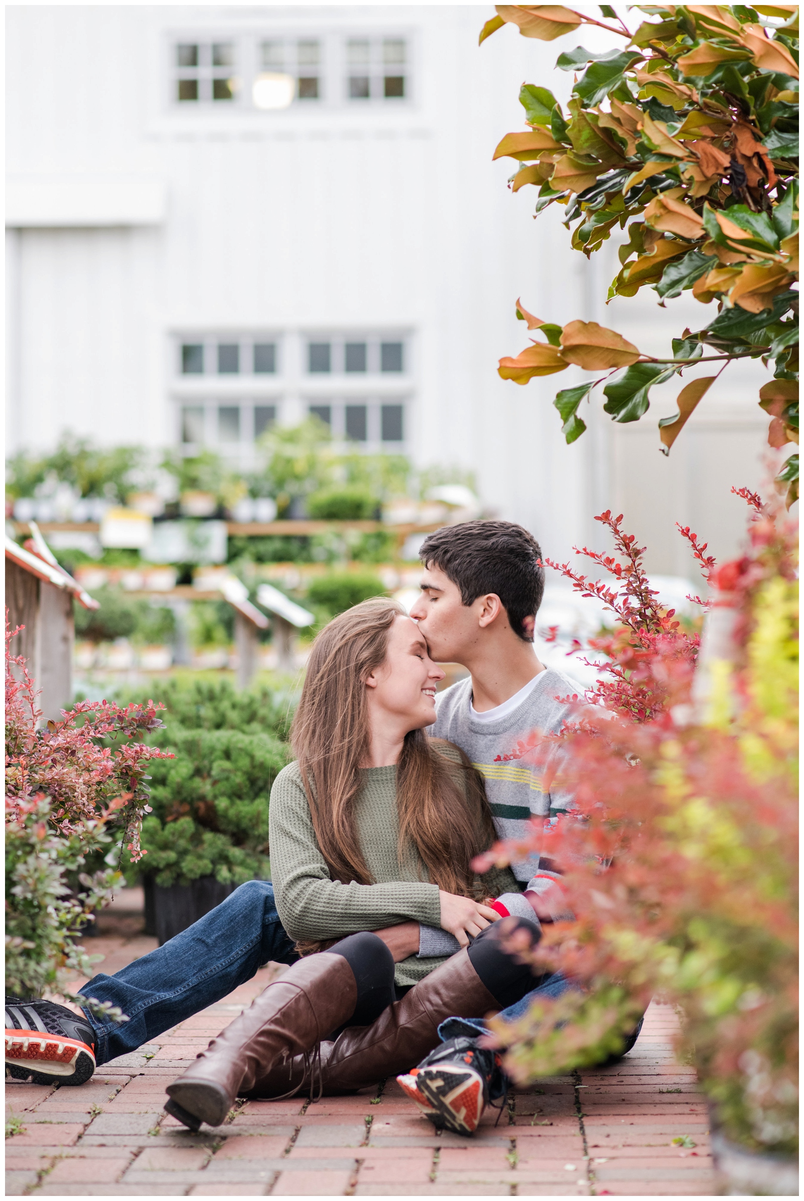 groom kisses bride on the forehead while they sit near red and orange plants during greenhouse inspired engagement session