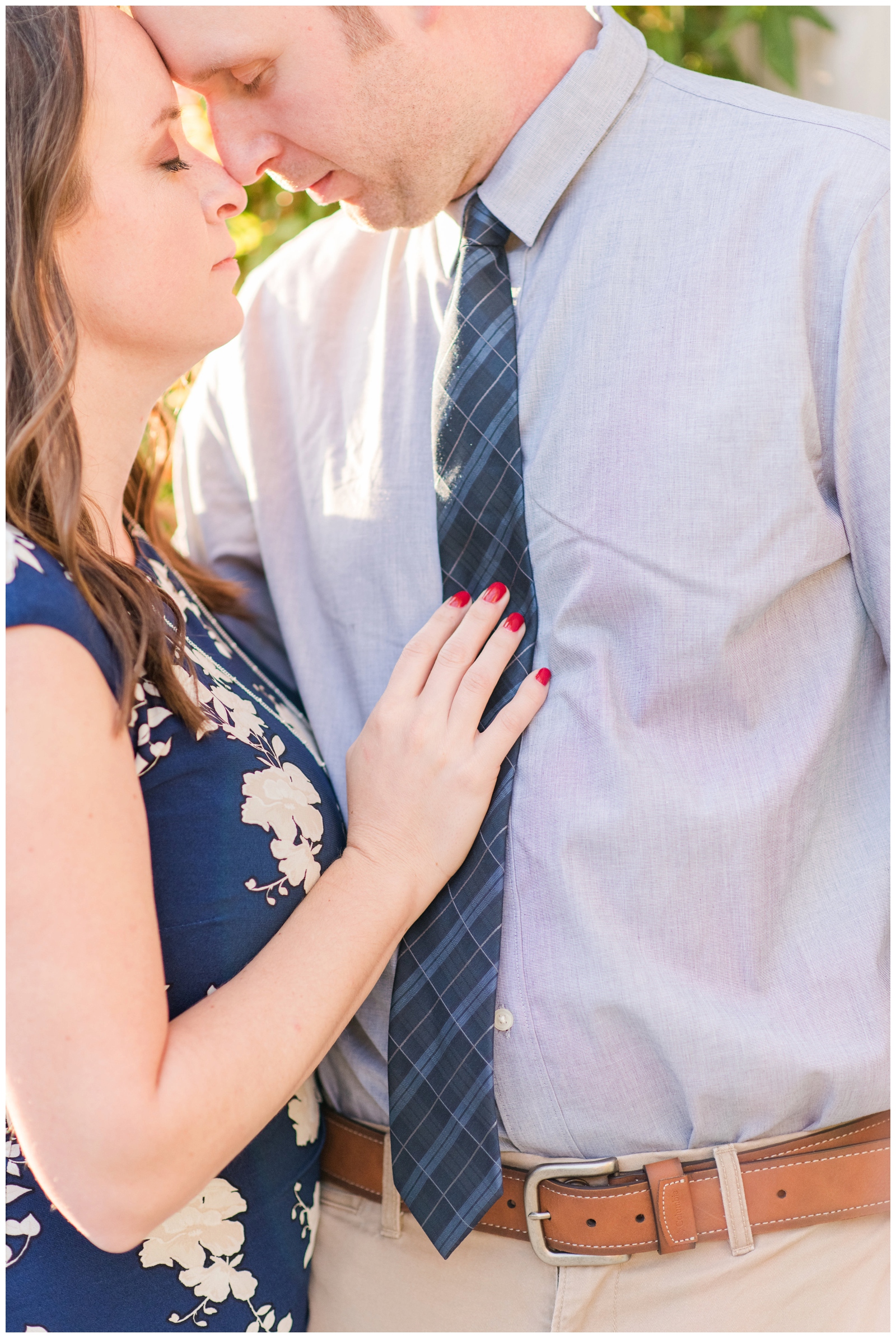 bride and groom touch noses during engagement portraits while bride touches groom's tie with red painted nails
