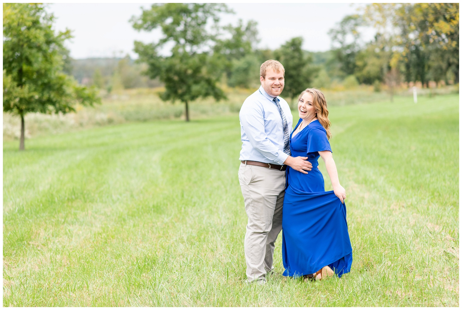 groom hugs bride while she twirls bright blue dress during summer engagement session Dawes Arboretum