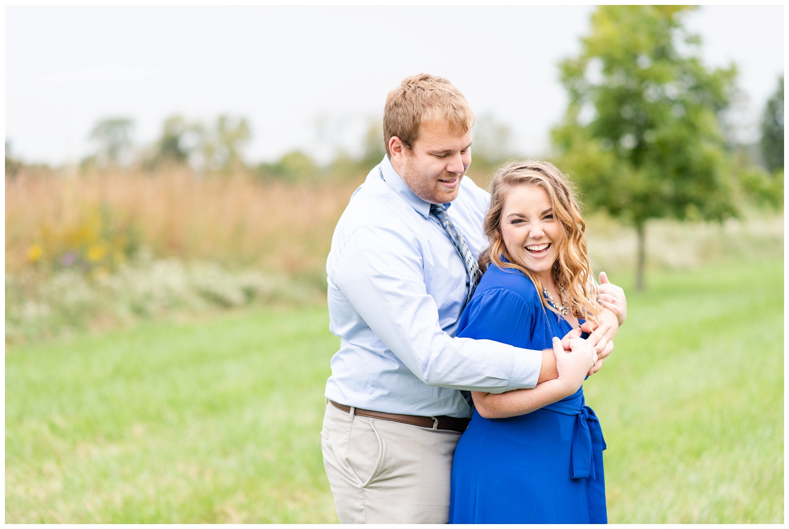 groom in blue button down shirt hugs bride in bright blue dress during summer engagement session Dawes Arboretum