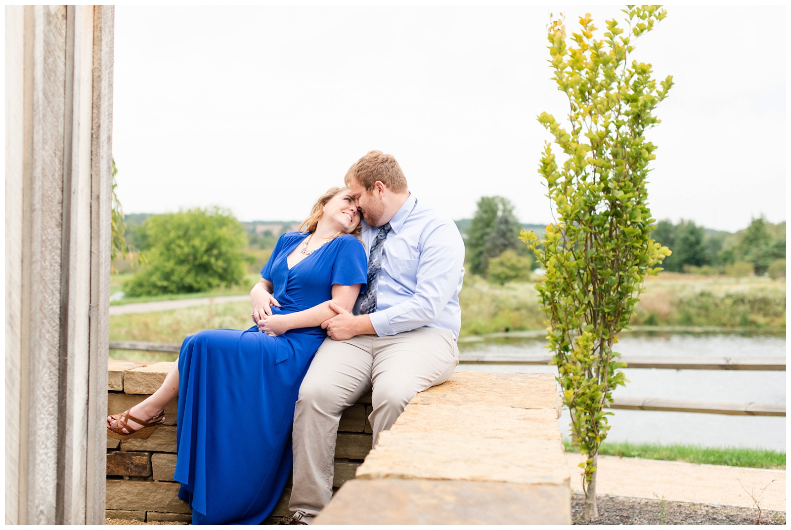 bride leans back against groom while sitting on brick wall and smiles