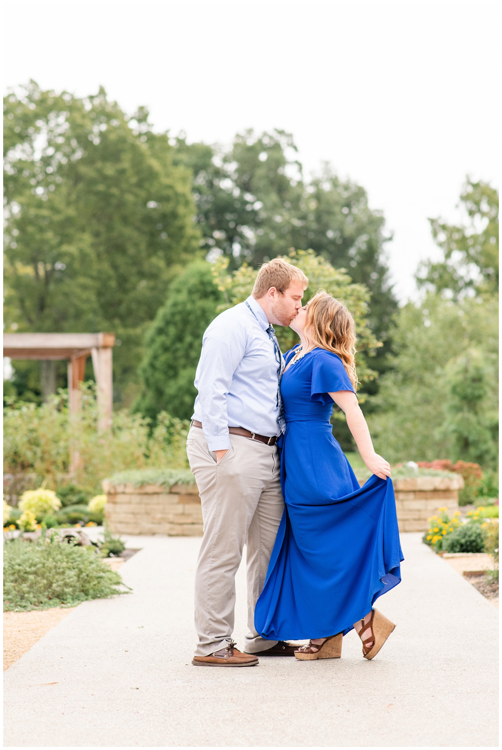 engaged couple kisses at Dawes Arboretum while bride-to-be holds out edge of bright blue dress