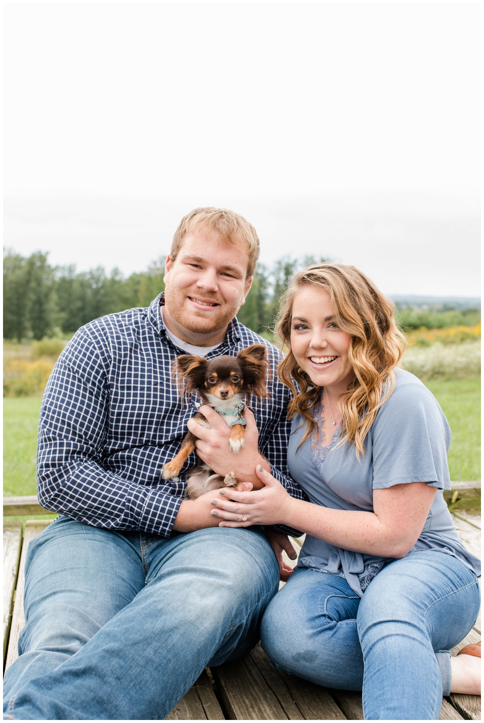 engaged couple snuggles with small dog while sitting on walkway at Dawes Arboretum