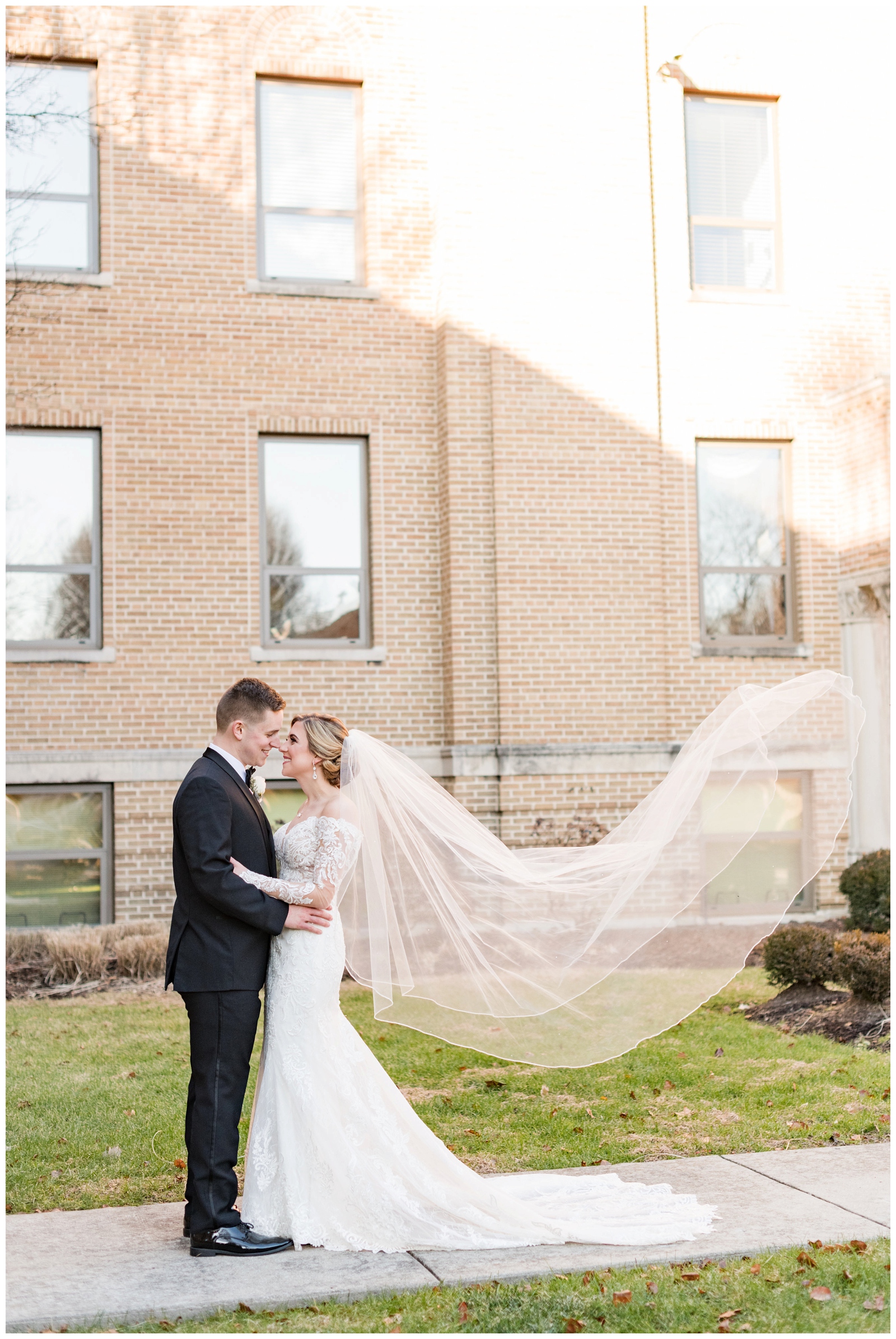 bride and groom stand nose to nose in front of St. Charles Preparatory School with veil floating behind bride