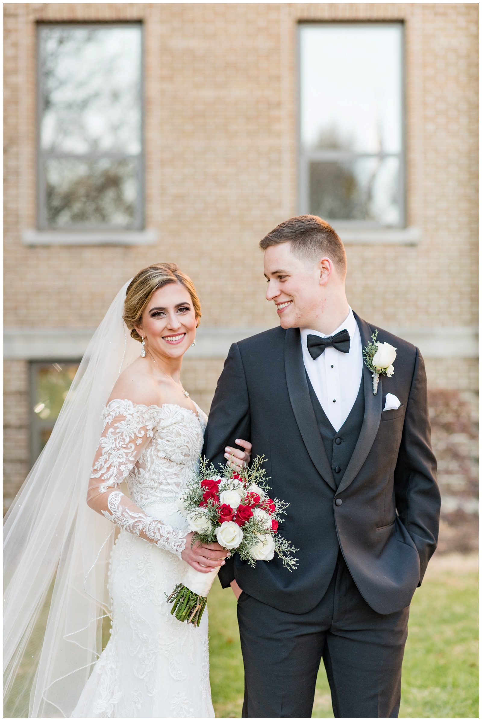 bride with long veil smiles at camera while groom looks at her in front of St. Charles Preparatory School