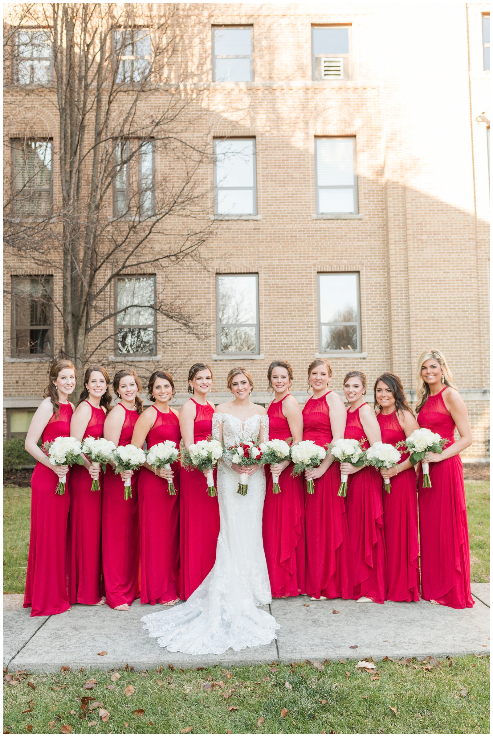 bride poses with bridesmaids in red gowns with white flowers for Christmas wedding
