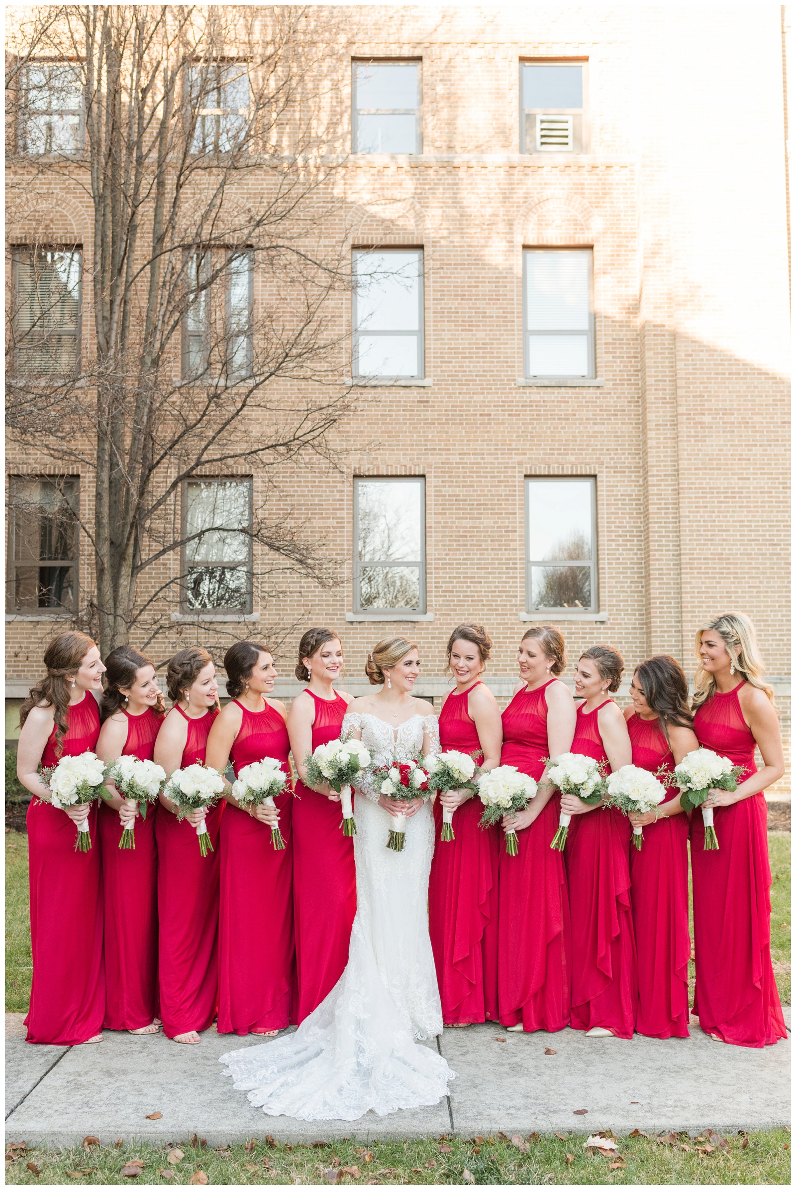 bridesmaids in red halter dresses with white rose bouquets look at bride smiling