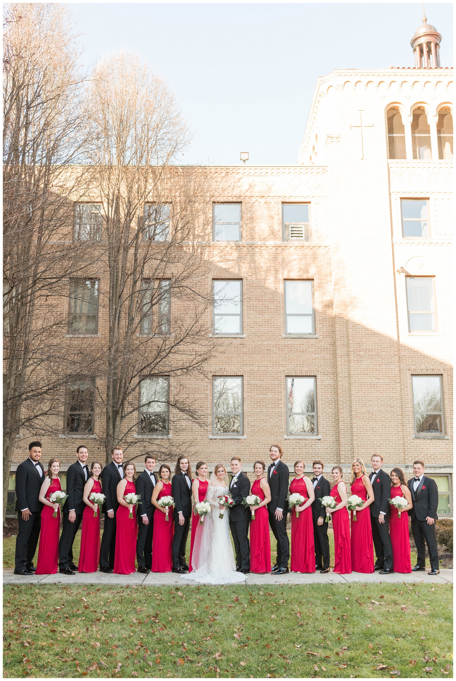 full wedding party in black tuxes and red dresses stand with bride and groom at St. Charles Preparatory School