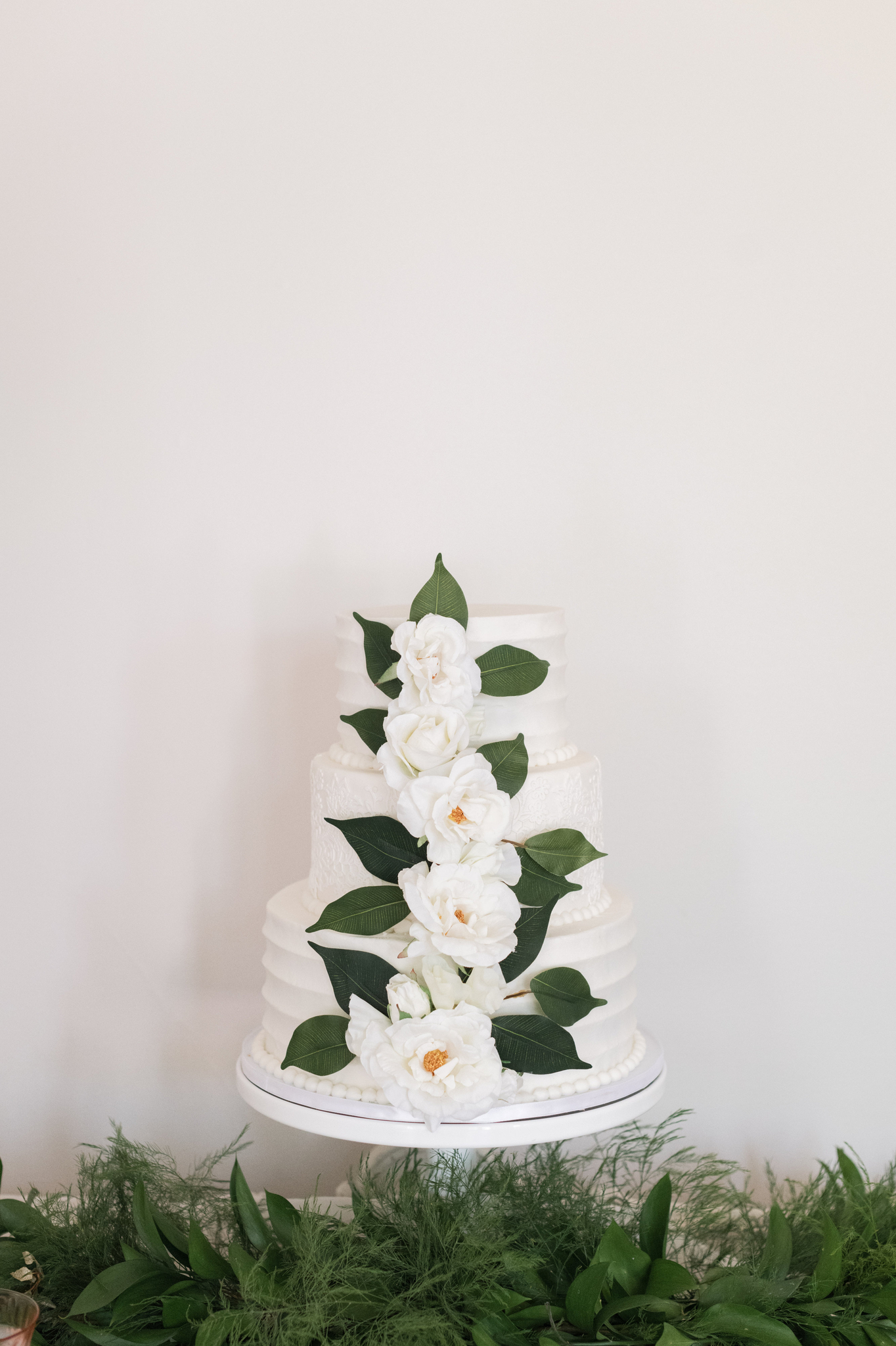 A three tier cake on a stand above greenery with white flowers on the front