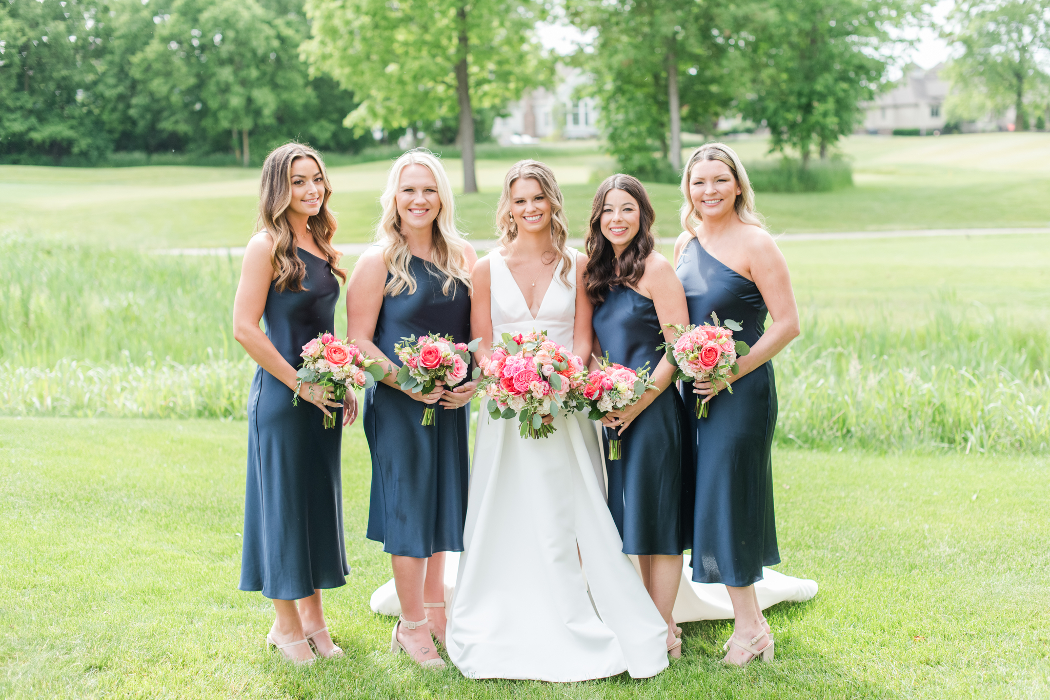 A bride smiles while standing with her bridesmaids in blue gowns holding pink bouquets