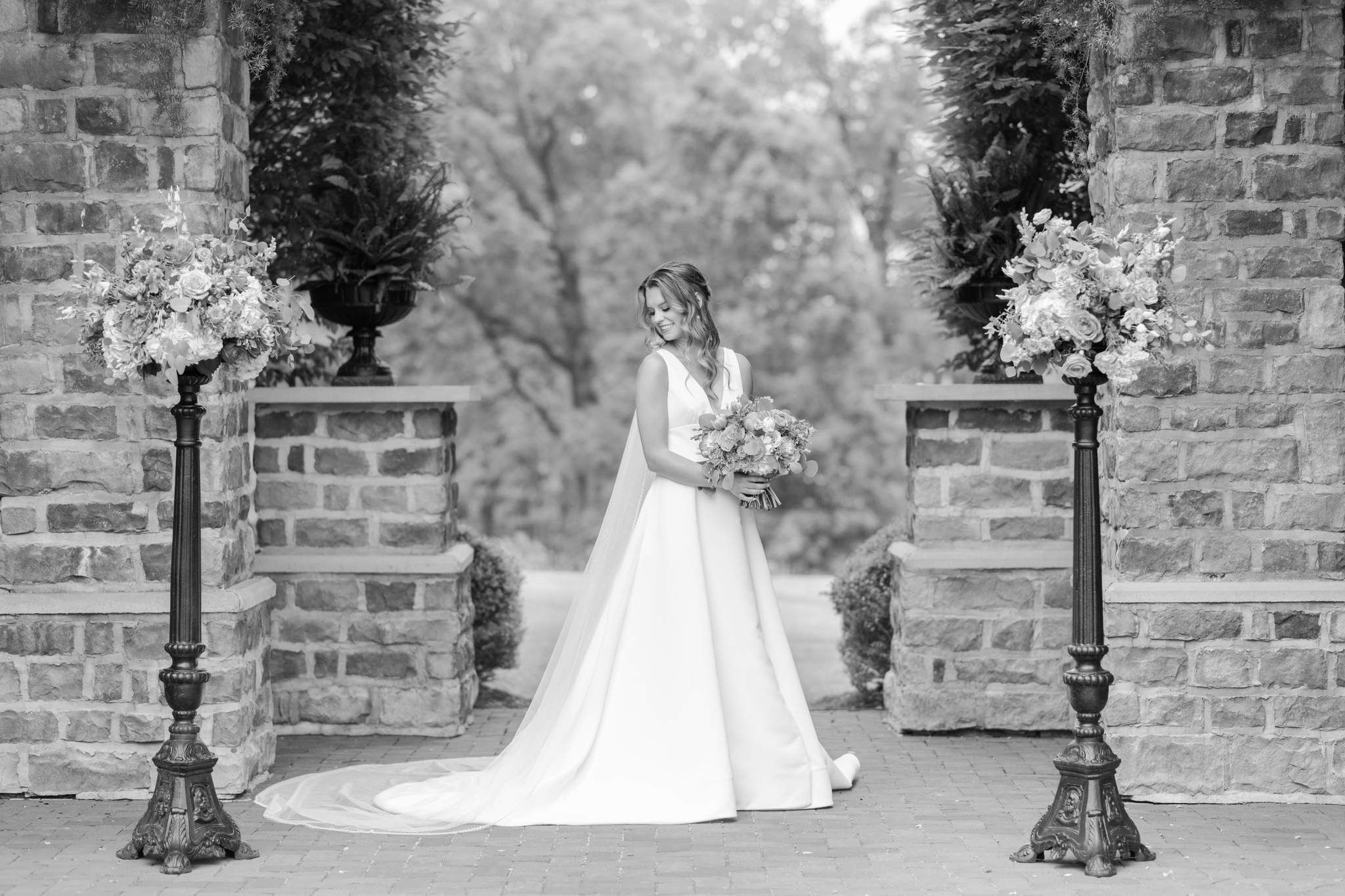 A bride smiles down her shoulder while standing in a large brick archway holding her bouquet at the Pinnacle Golf Club Wedding venue in black and white