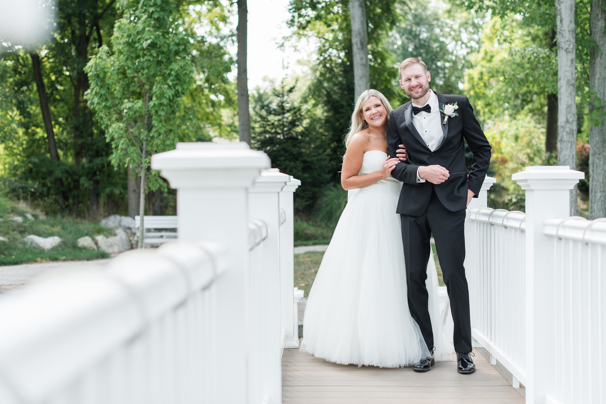 A bride hangs on the arm of her groom as they cross a white bridge over a pond