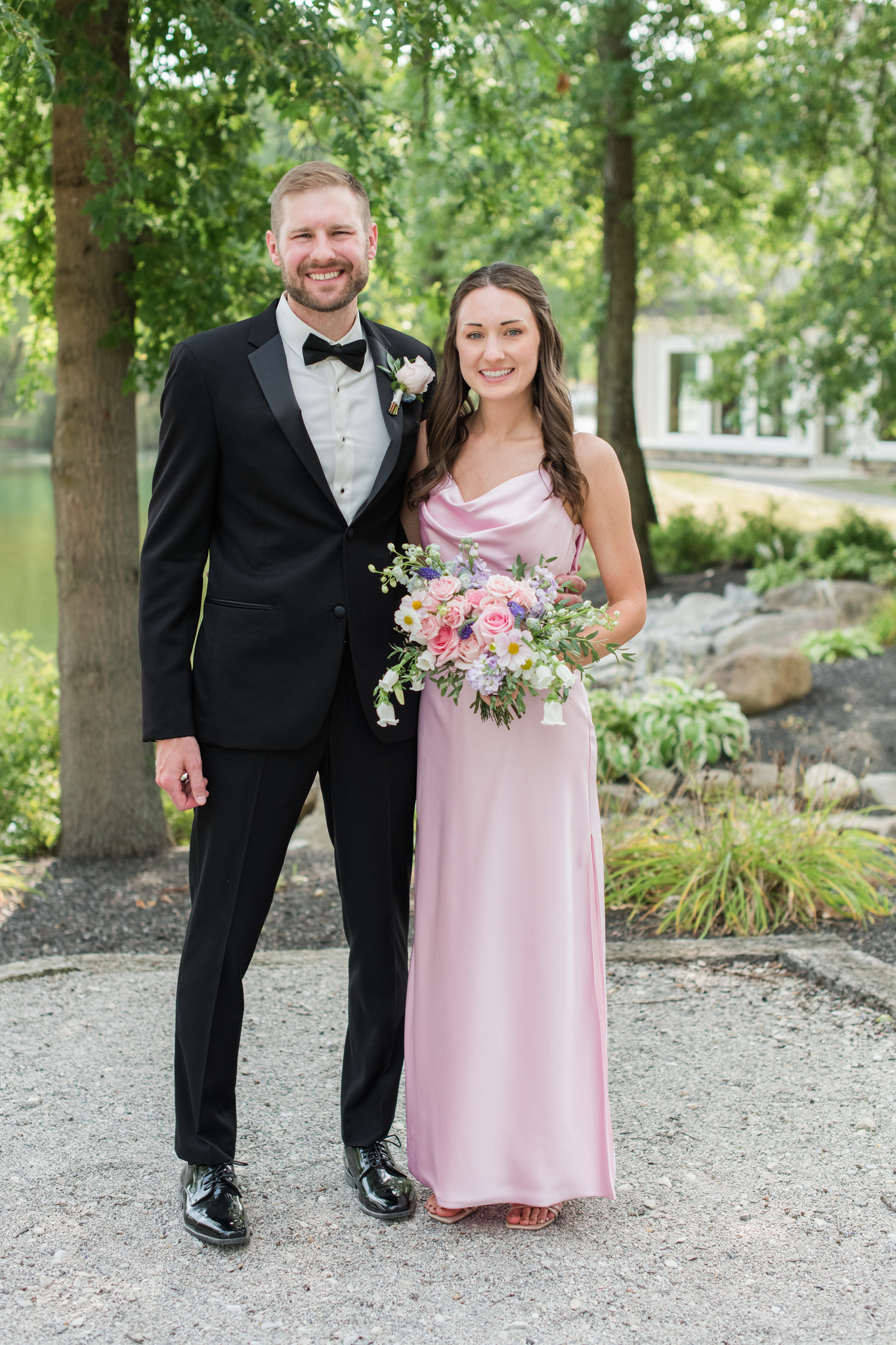 A groom smiles with a bridesmaid in a pink dress and colorful bouquet in the gardens of the Swan Lake Event Center