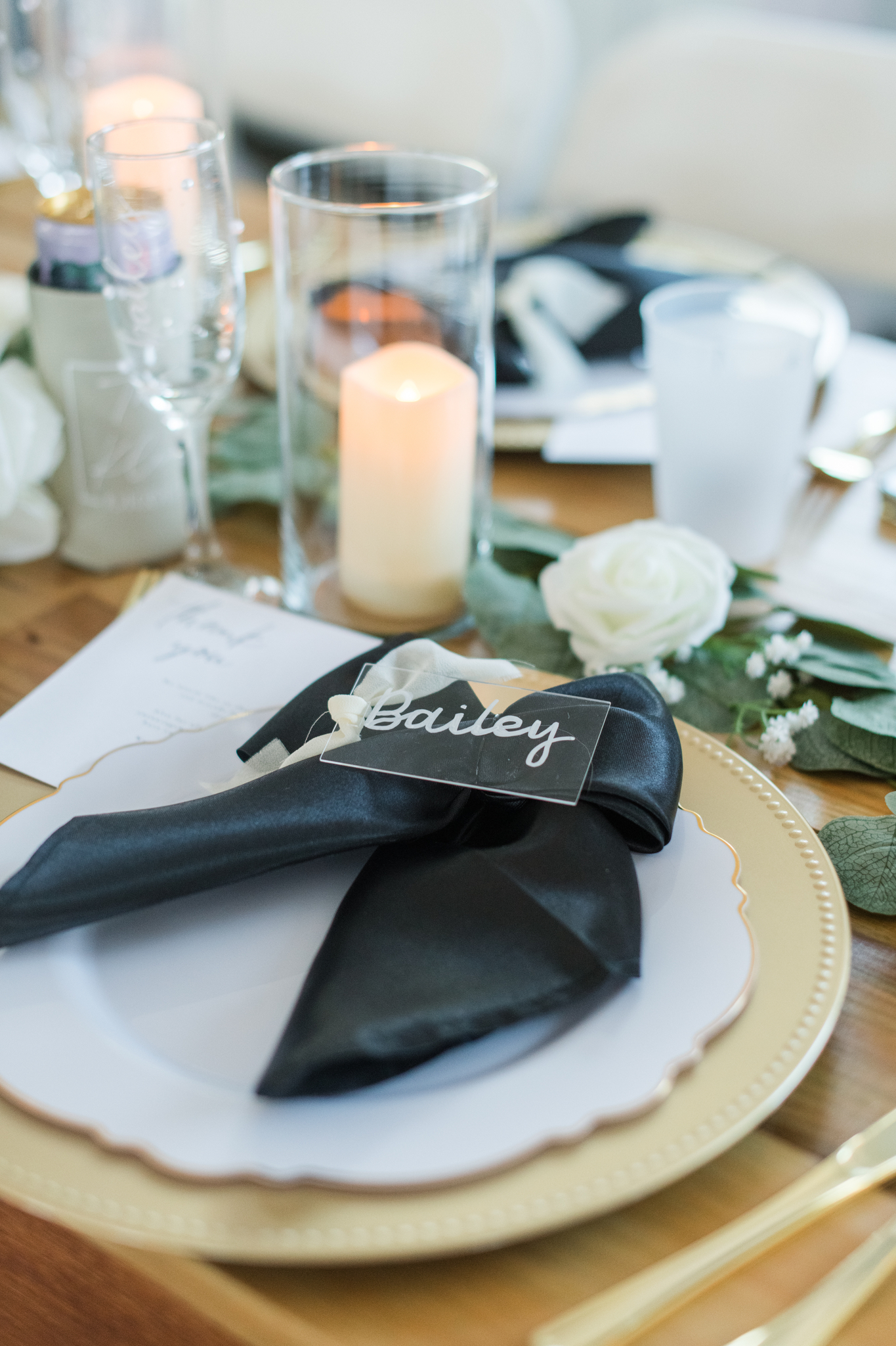 A custom name plate rests on a black napkin of a White Rose Barn wedding reception place setting with white roses